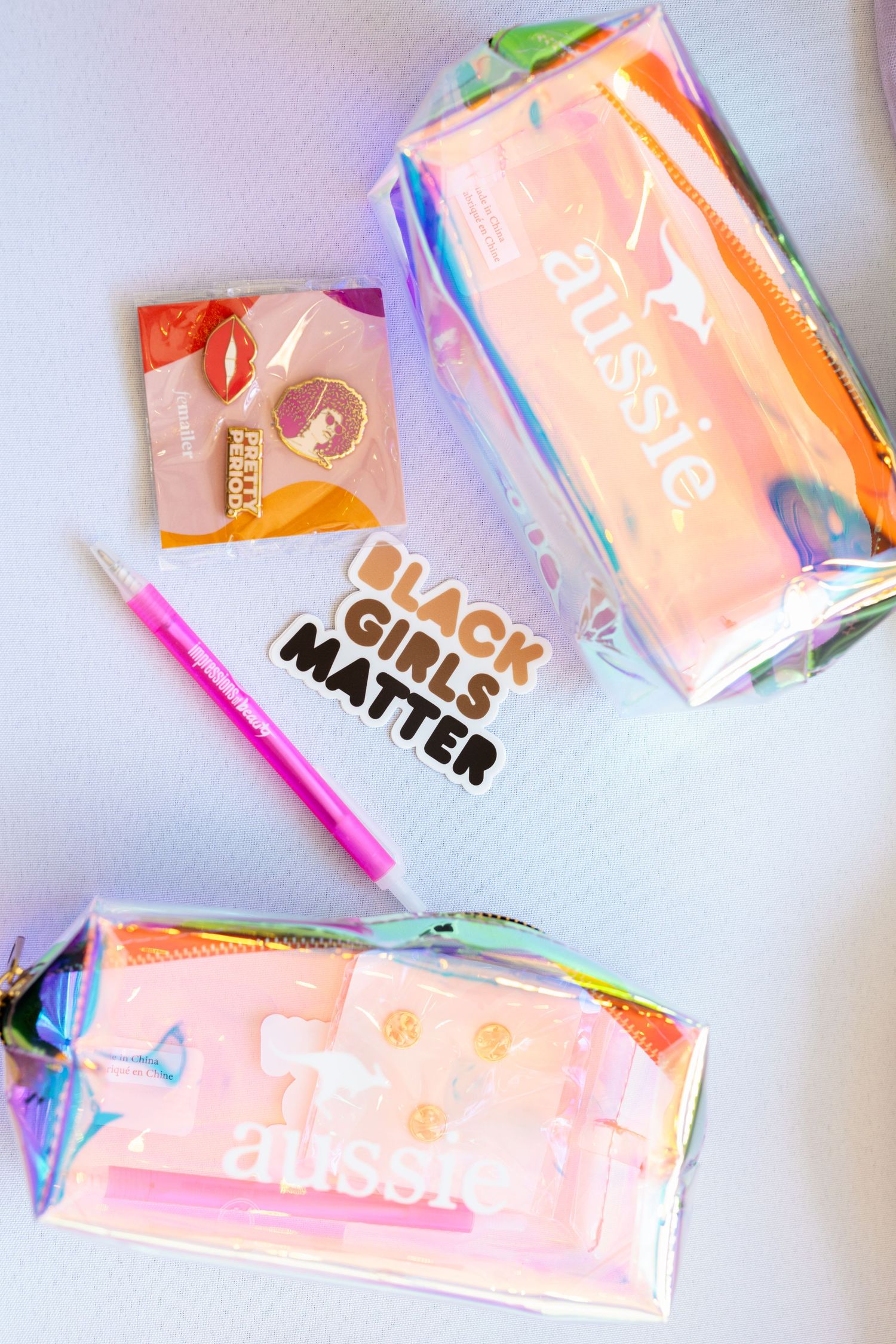 Two iridescent Aussie hair product bags with a Black Girls Matter sticker, pink pen, and beauty samples on white surface.