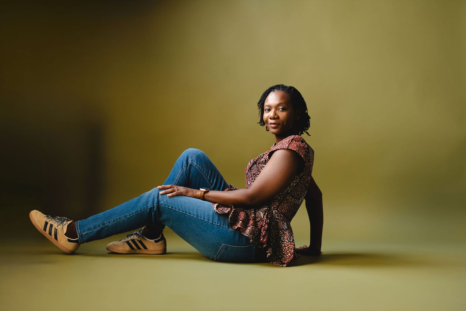 Young woman in jeans and floral top sitting relaxed on floor, smiling confidently against yellow-green backdrop.