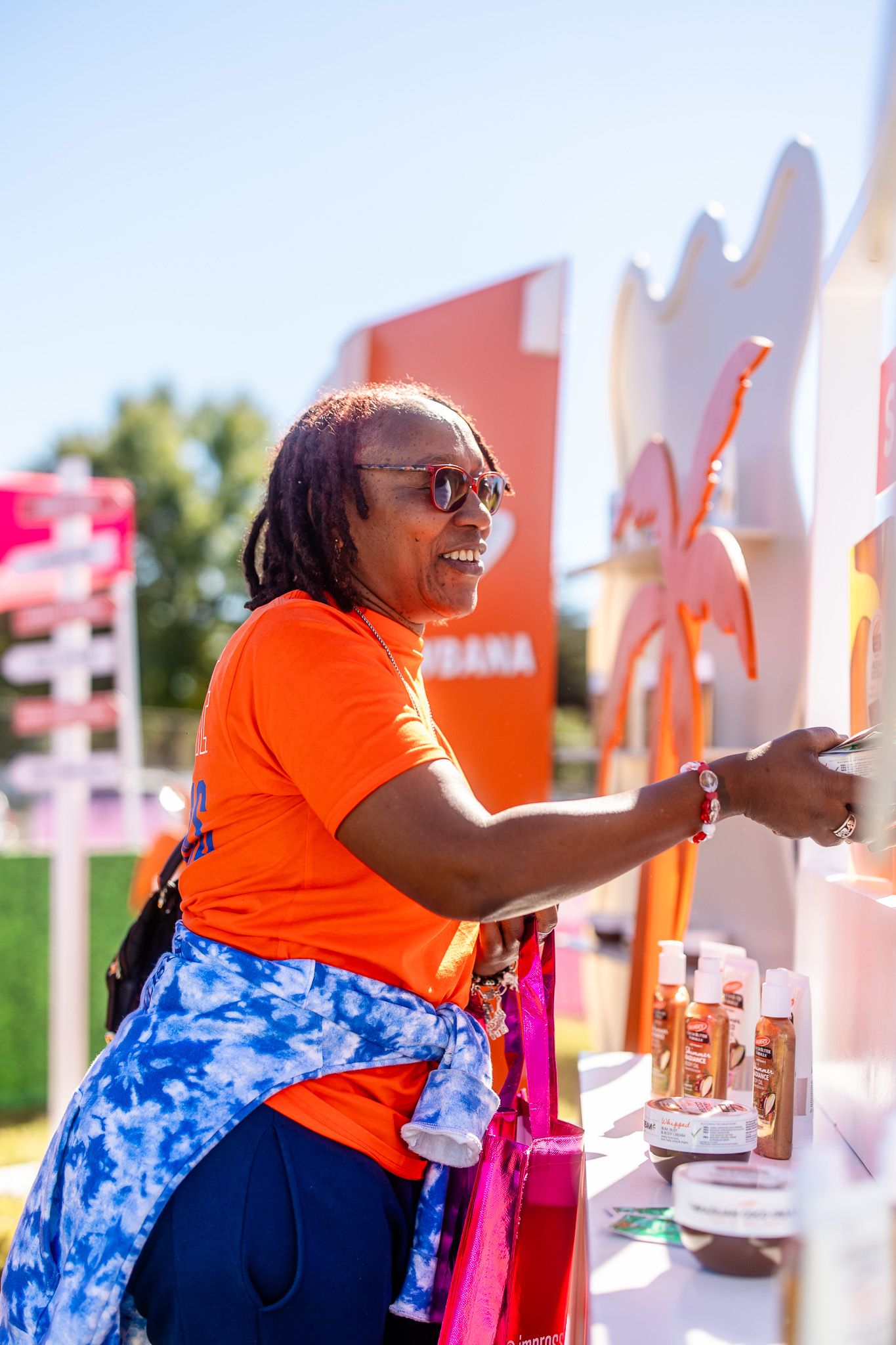 Woman in orange shirt smiling at outdoor product display booth on sunny day.