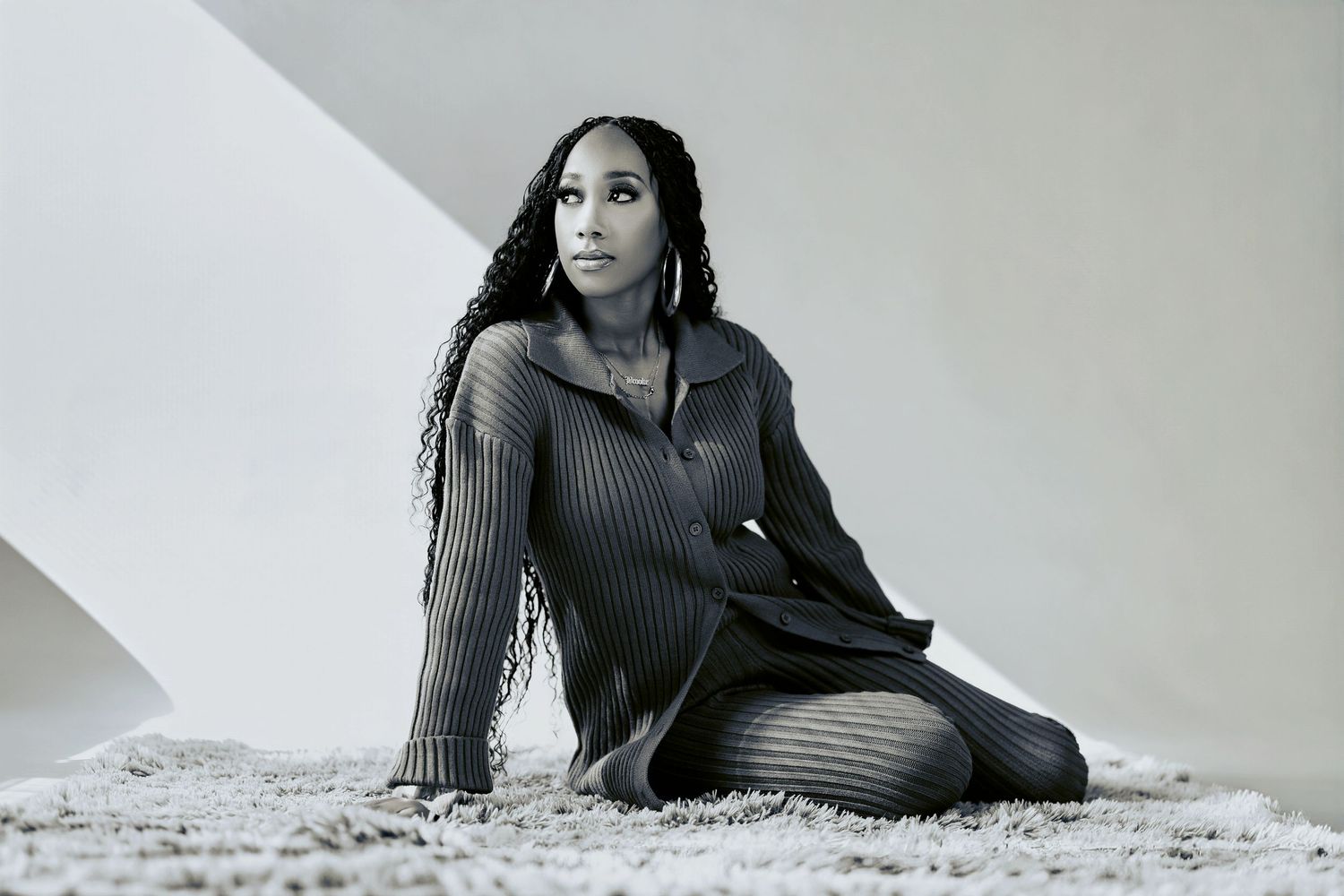 A woman with long braids sits gracefully on a carpet in a ribbed knit dress, posed against geometric white backdrop.