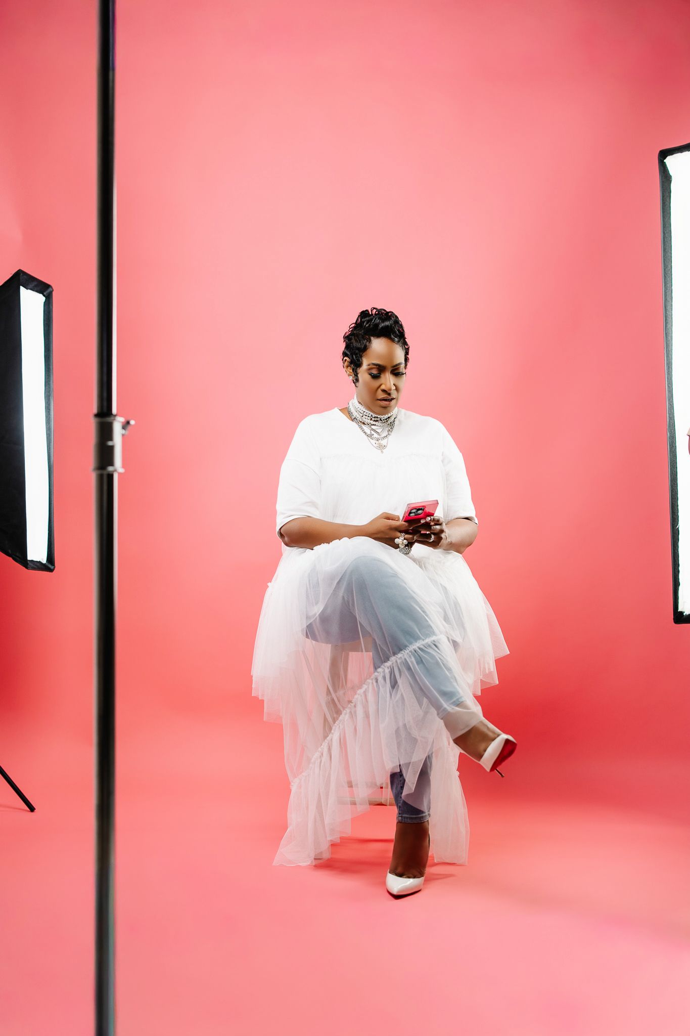 Woman in white tulle skirt uses phone in pink photography studio with professional lighting equipment.