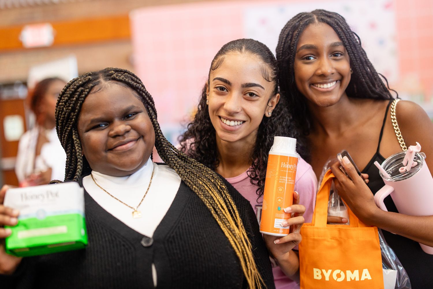 Three young women smiling and posing together, holding BYOMA skincare products at a beauty event.