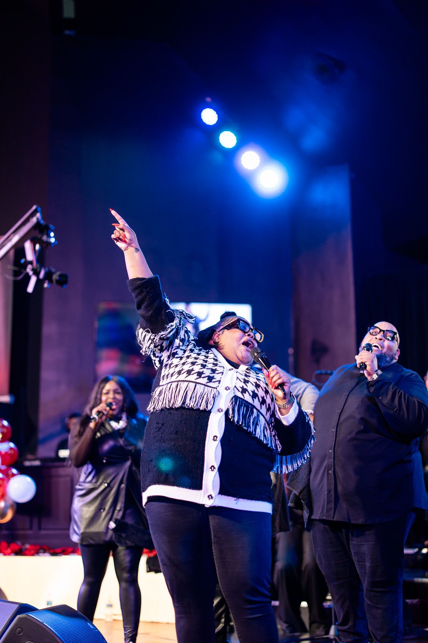 Gospel singers performing on stage with dramatic blue lighting, one singer pointing upward with microphone at live concert.