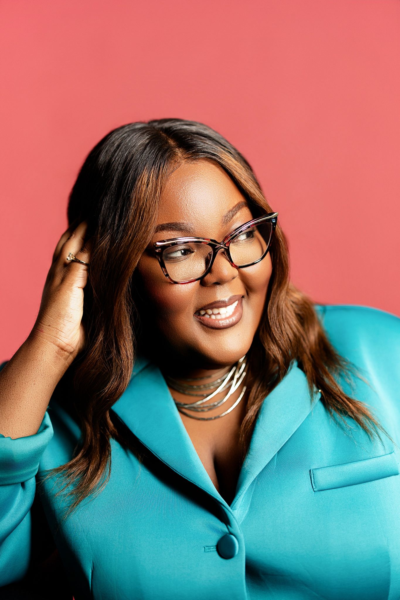 Smiling woman in teal blazer and tortoise shell glasses poses against a vibrant pink background.