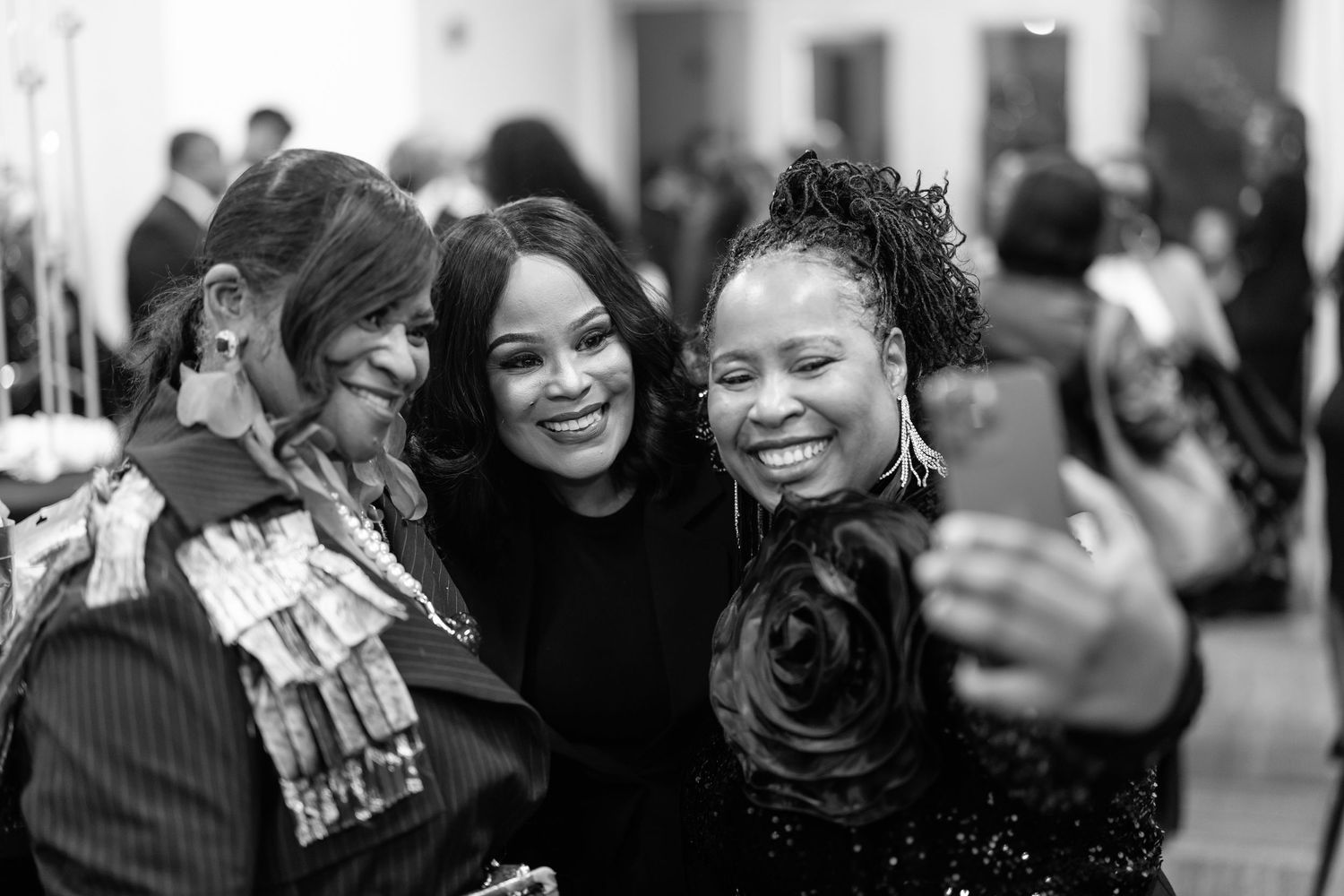 Three women smile together for a selfie at a formal event in this black and white photo.