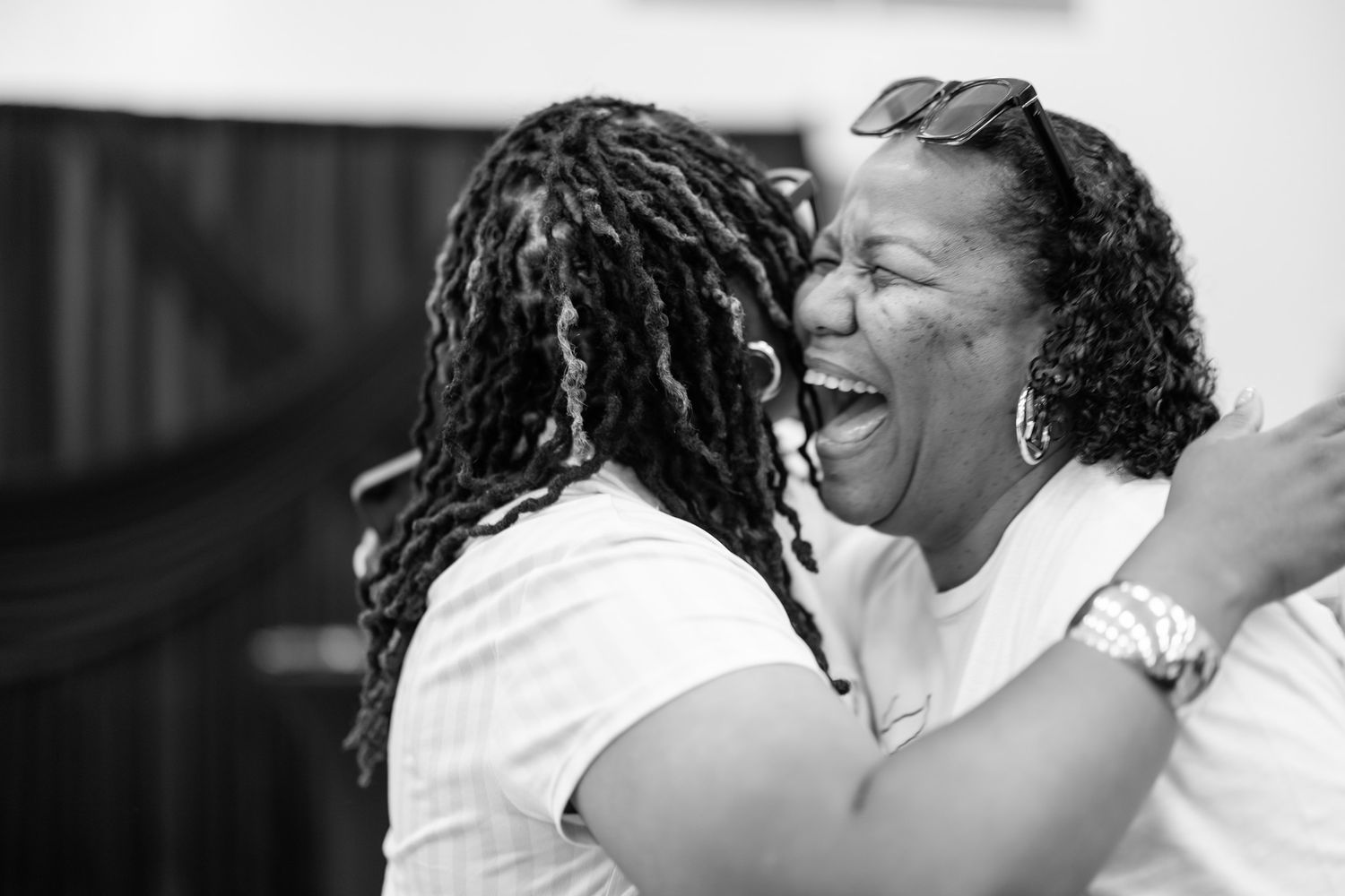 Two Black women share a joyful embrace and laugh together in a warm black and white photo.