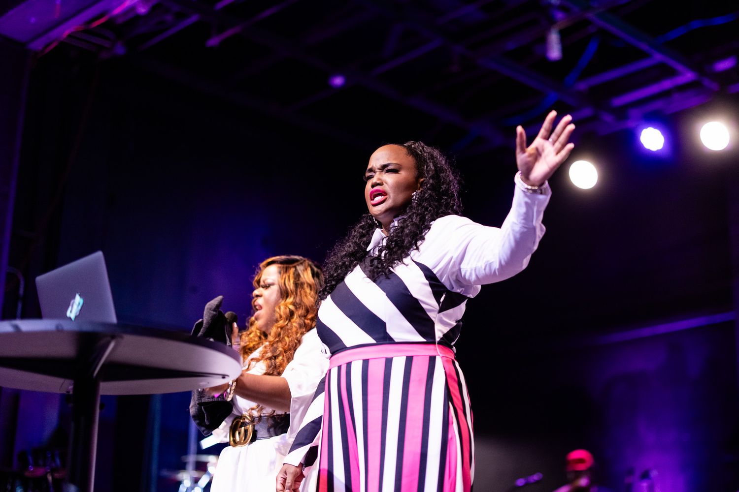 Two women performing on stage, one gesturing expressively, wearing striped outfits under colorful stage lights.
