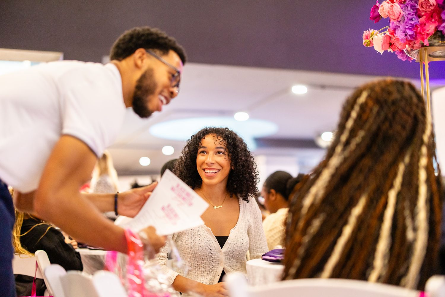 Man and woman smiling at a elegant event, she holds pink papers while another woman with braids sits nearby.