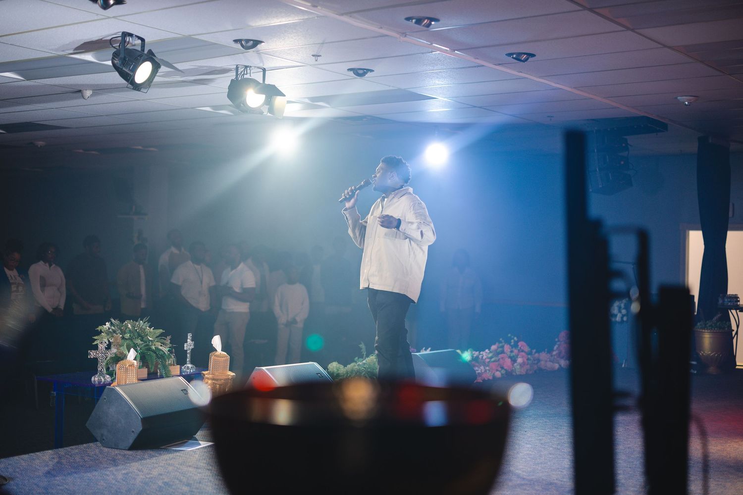 Performer on stage with microphone under blue stage lights at an indoor venue with audience watching.