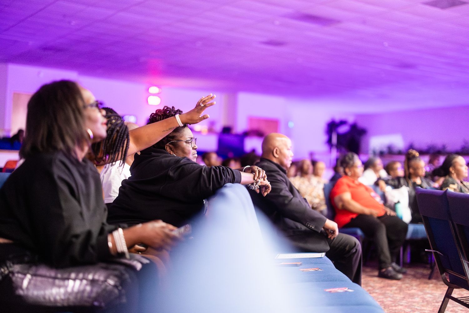 Audience members seated in a purple-lit event hall, with one person raising their hand during a conference.