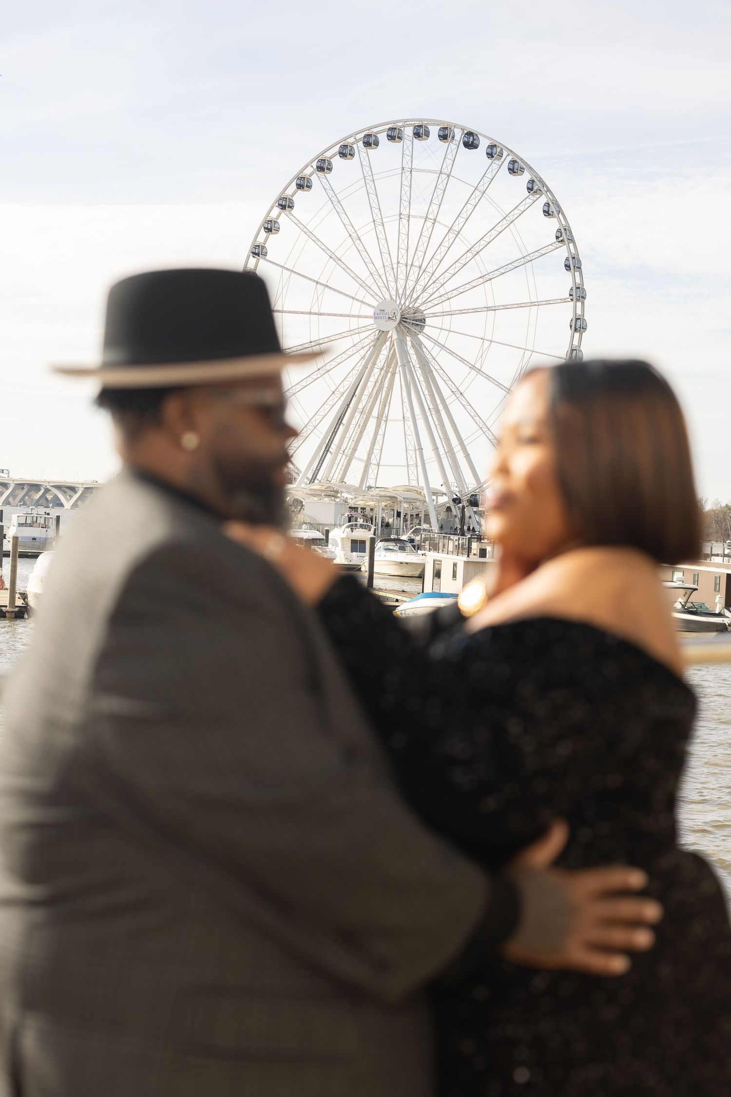 Couple embracing at National Harbor waterfront with iconic Capital Wheel Ferris wheel in background.