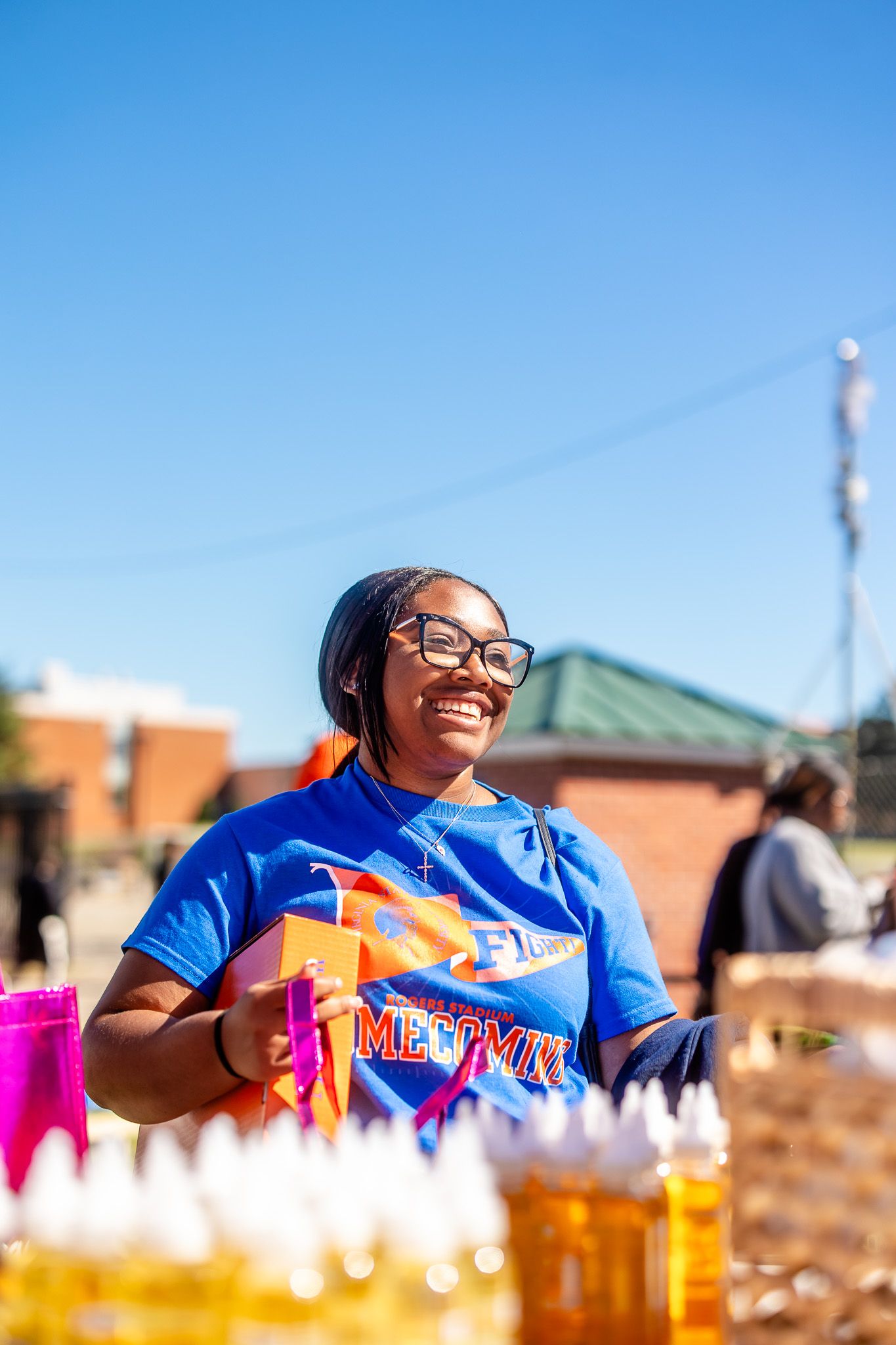 Smiling young woman in blue Homecoming shirt holds gift at sunny outdoor event with colorful bottles in foreground.