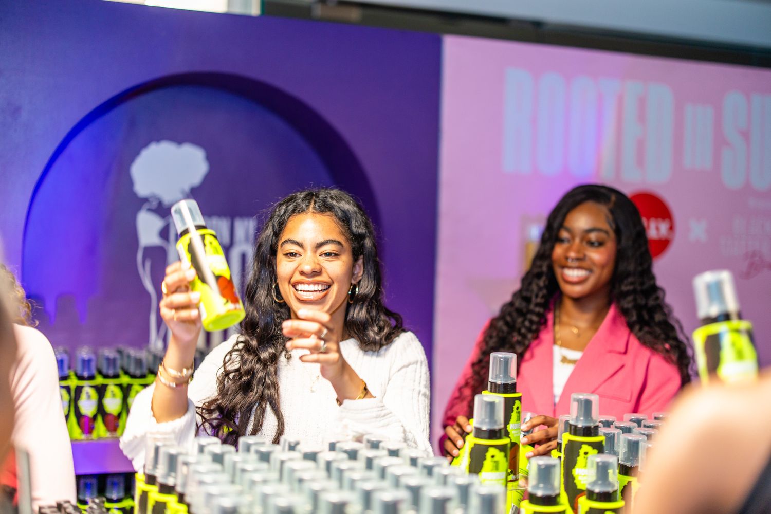 Two women smile at a product launch event, one holding up a green bottle among rows of displayed products.
