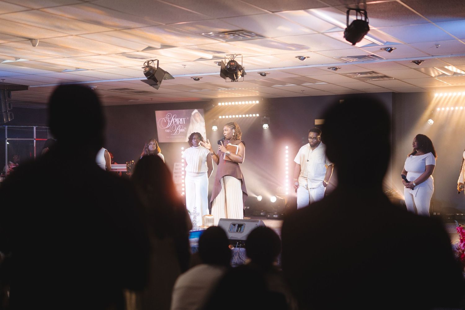 Performers in white outfits on a lit stage with silhouetted audience members watching during a live event.