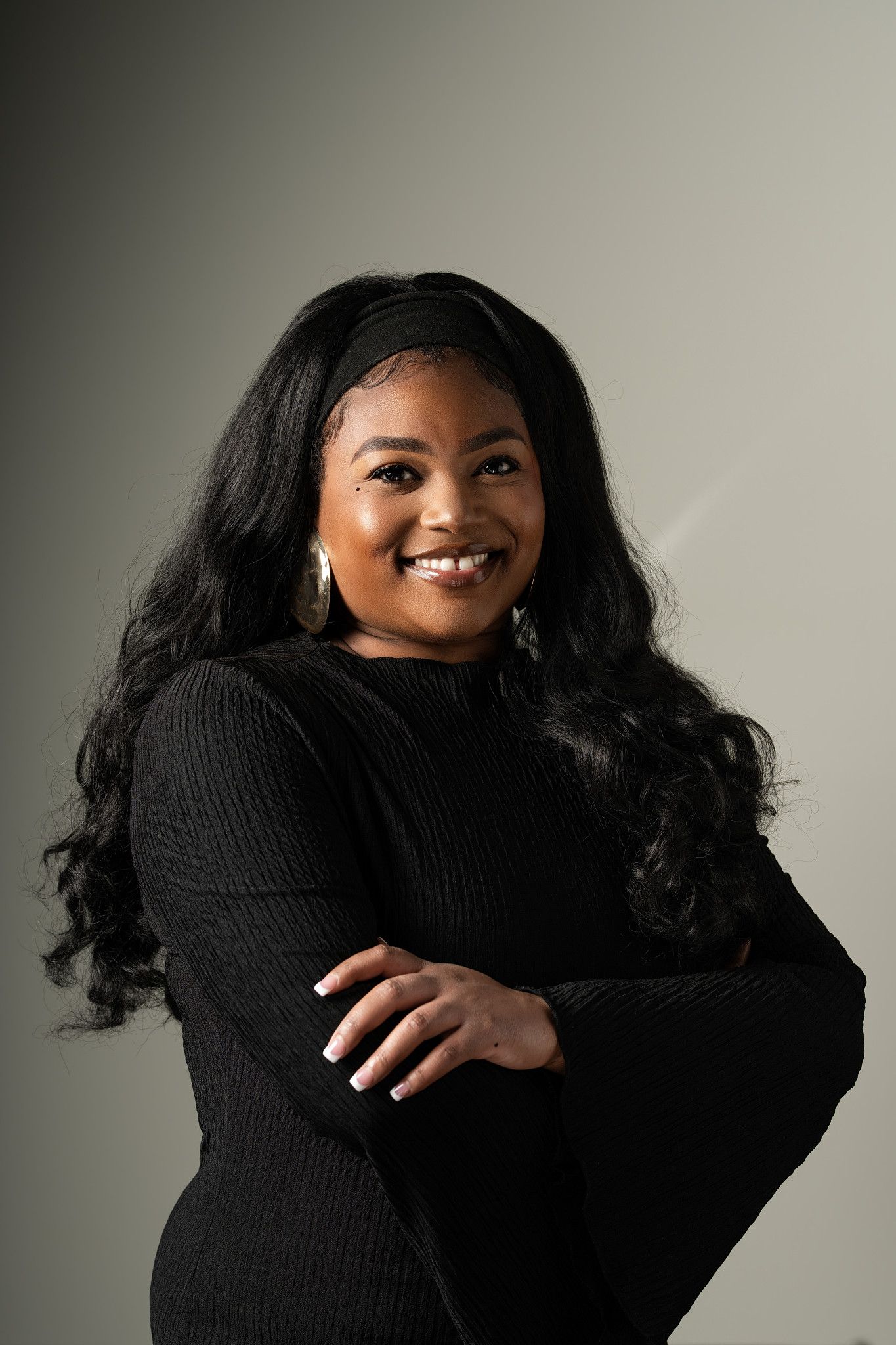 Smiling Black woman with long wavy hair wearing a black ribbed sweater, posing with arms crossed against a gray background.