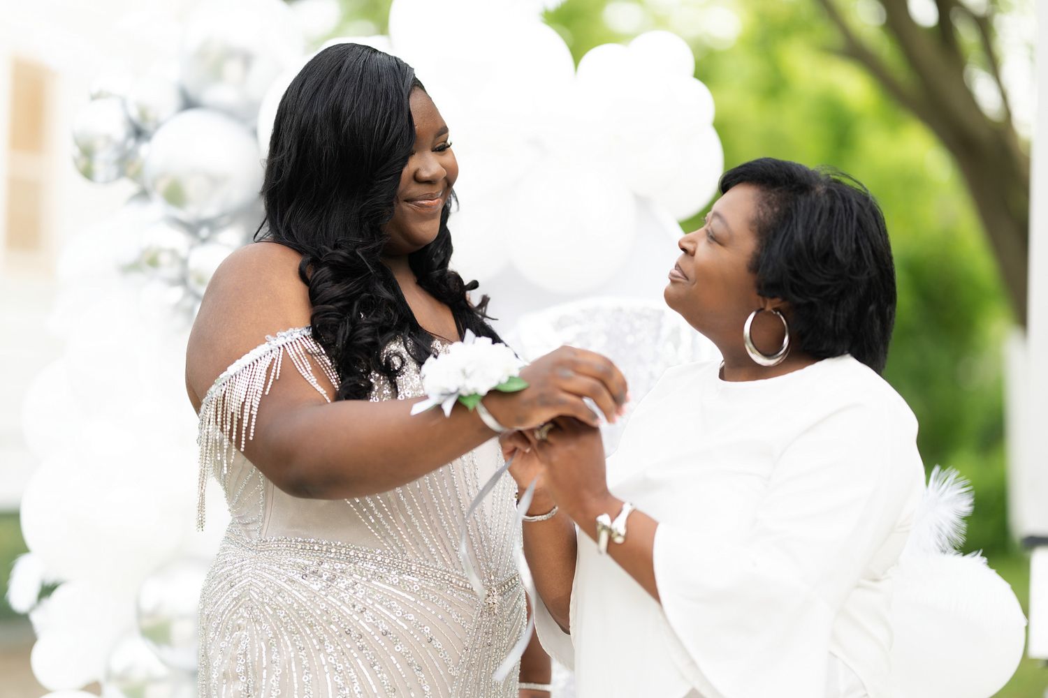 Two women in white attire share a joyful moment outdoors surrounded by white balloons at a celebration.