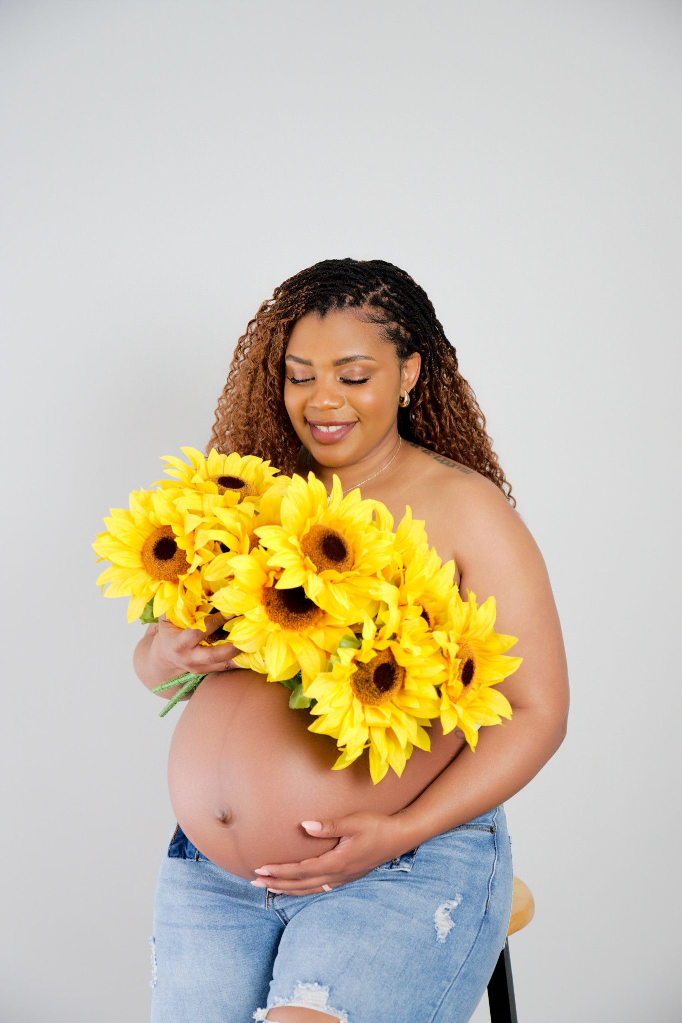 Pregnant Black woman with curly hair holds bright yellow sunflowers, smiling down at her bare baby bump in jeans.
