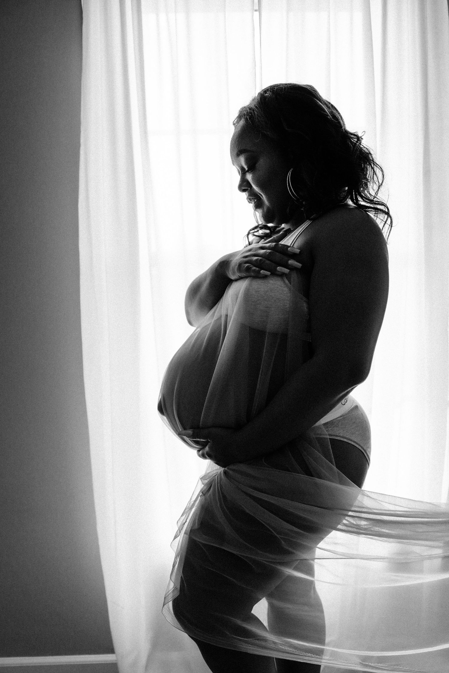 Silhouette of pregnant woman wrapped in sheer fabric standing by a bright window in black and white.