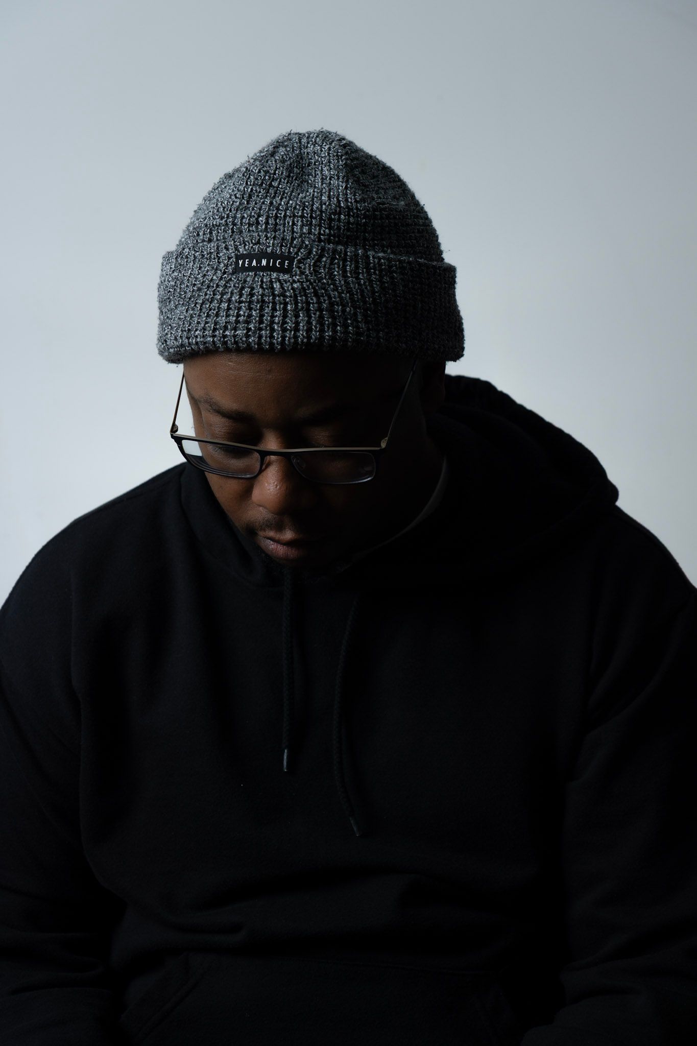 Young Black man in black hoodie and gray beanie looking downward against white background.