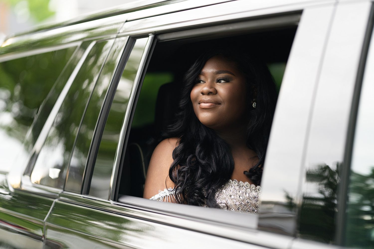 A smiling young woman in a beaded gown looks out a limousine window, dressed elegantly for prom or a special event.
