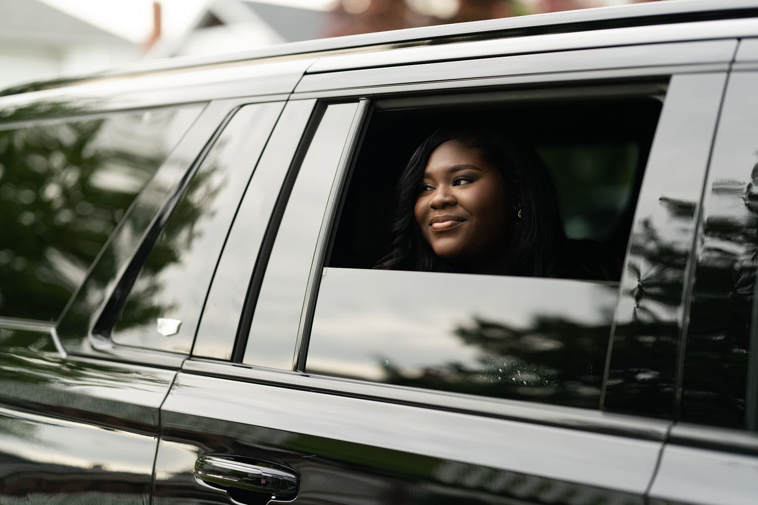 A smiling young Black woman looks out the window of a sleek dark SUV or limousine.