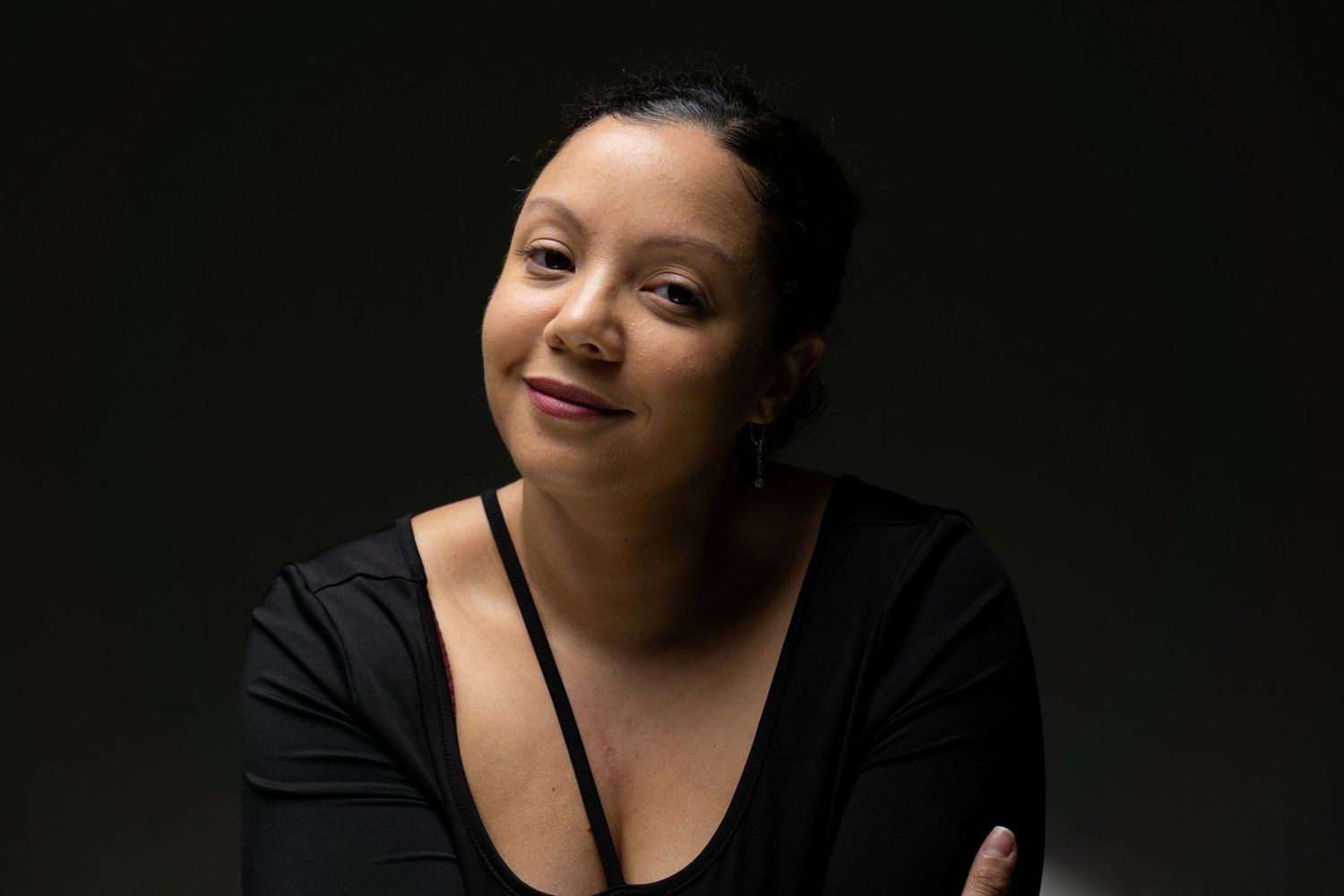 Woman with crossed arms smiling softly in a black top against a dark studio background.