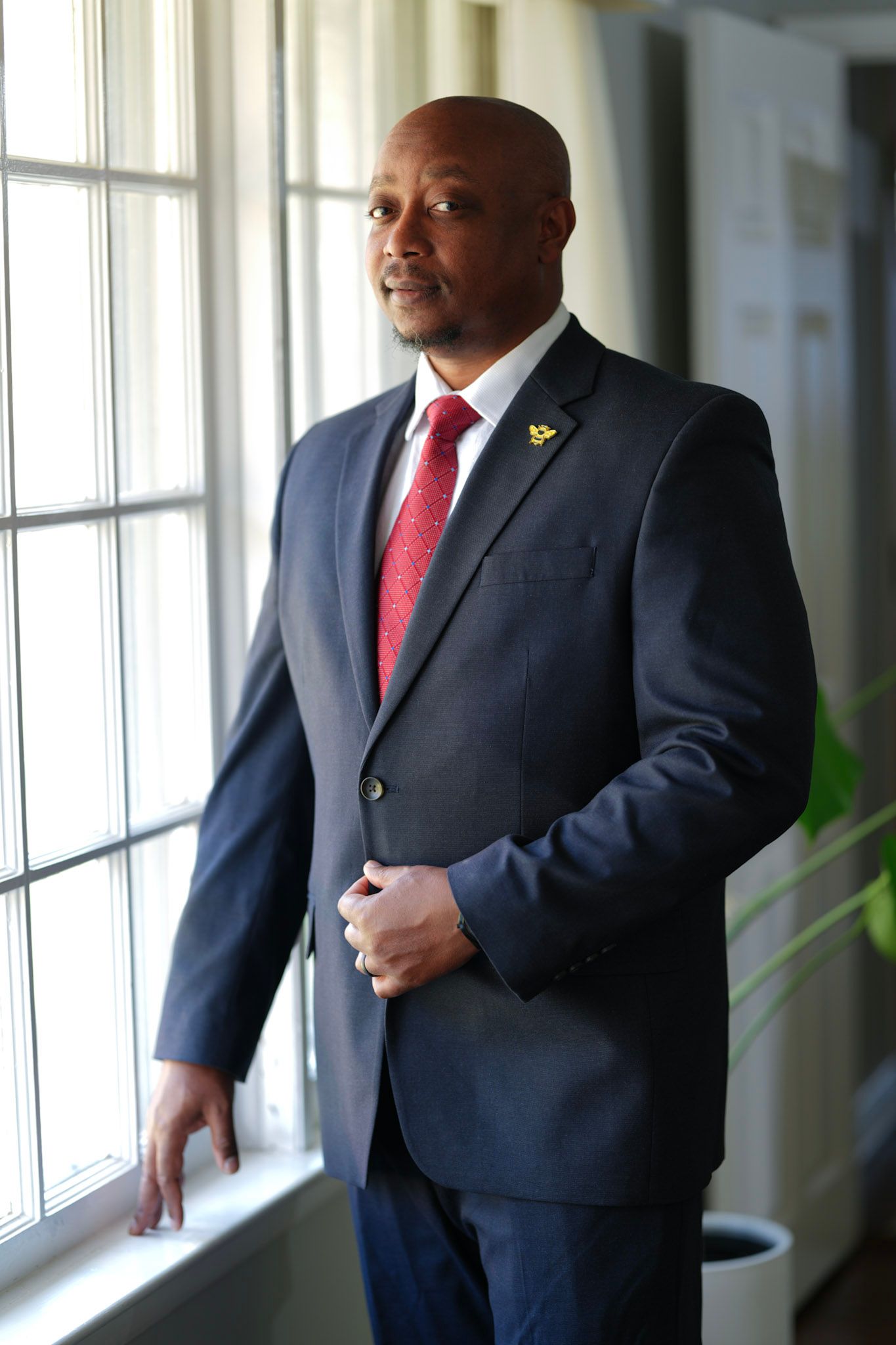 Confident Black businessman in navy suit and red tie standing by a sunlit window.