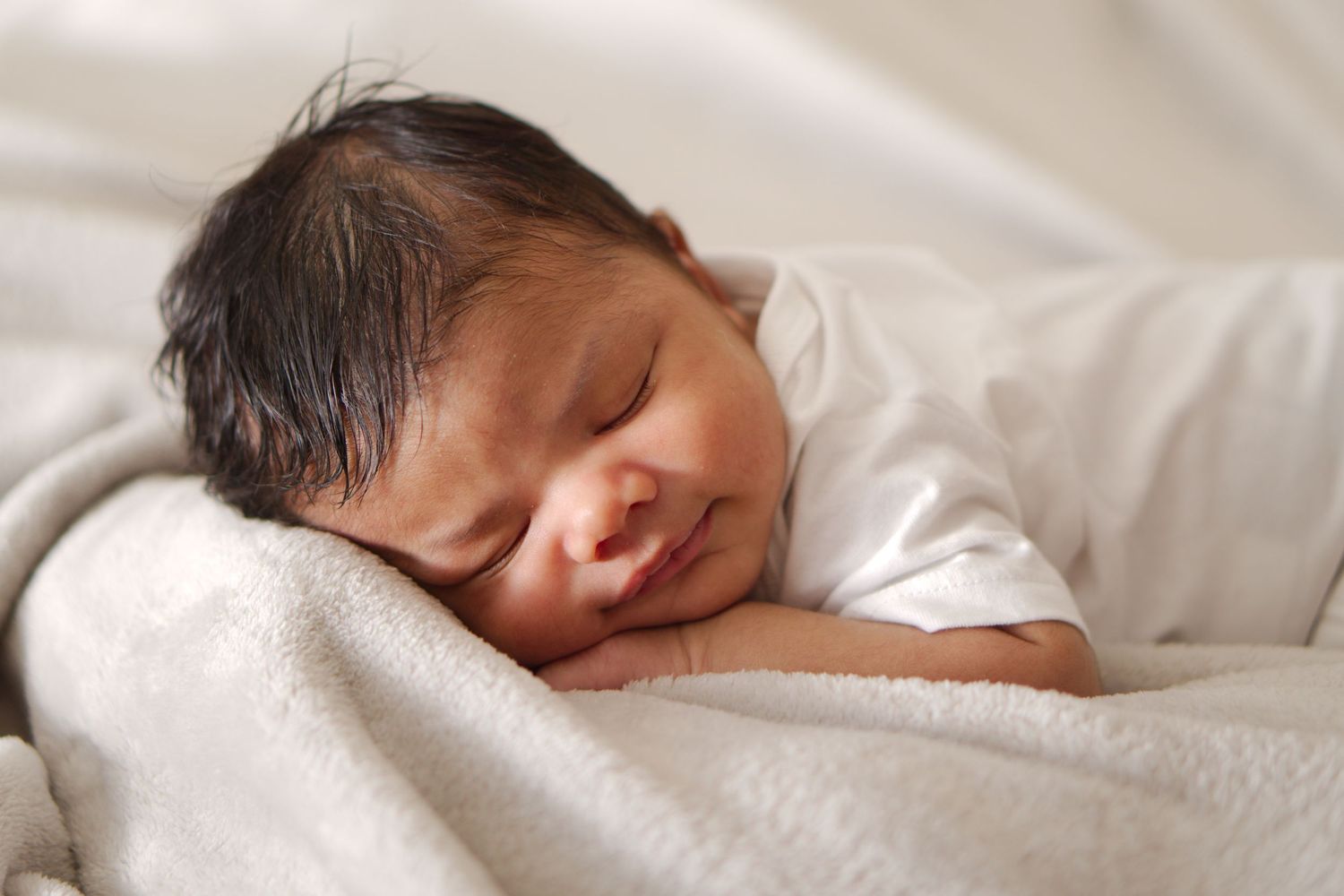 Peaceful sleeping newborn baby with dark hair resting on a soft white blanket.