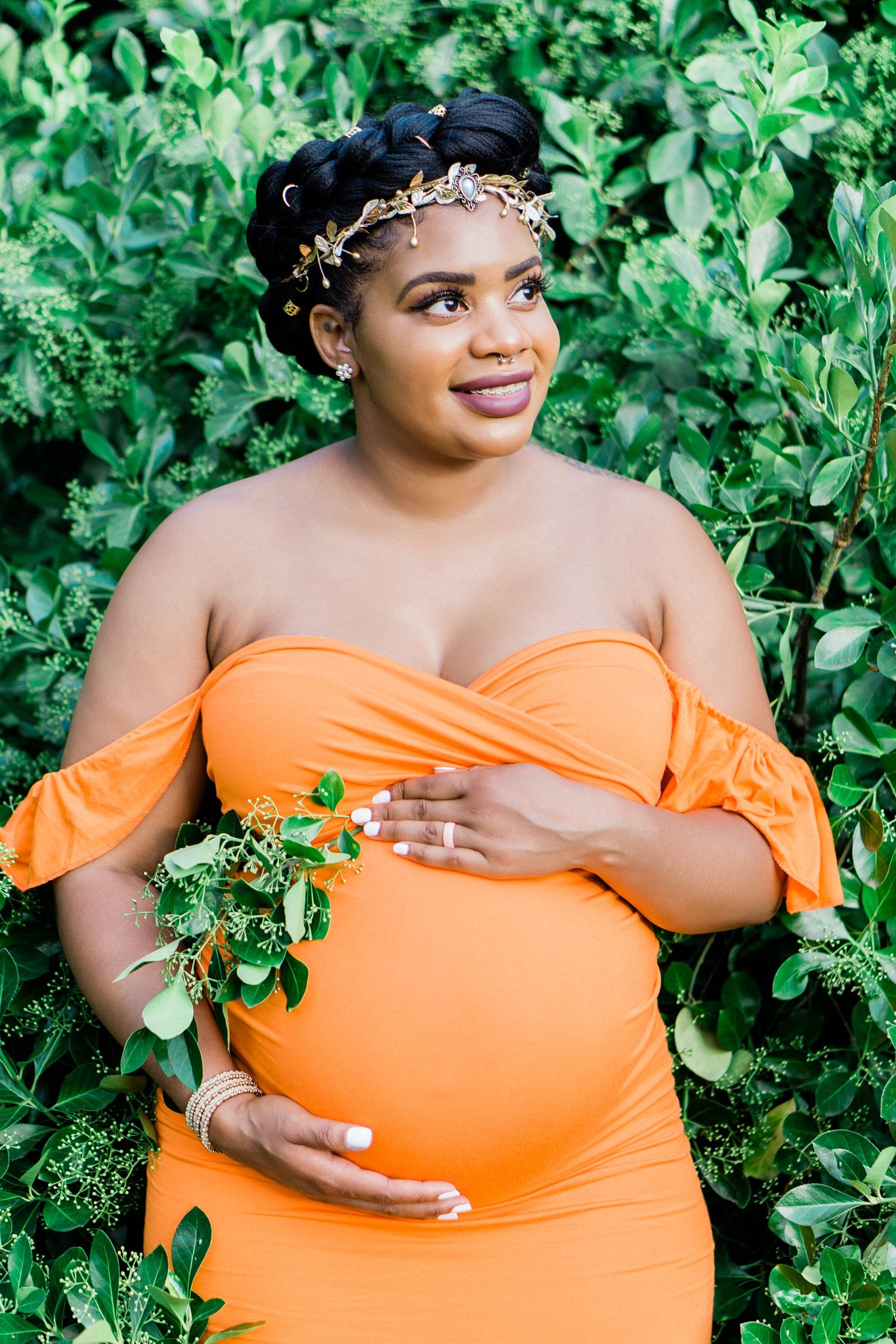 Pregnant woman in orange off-shoulder dress with gold crown poses gracefully among lush green foliage.