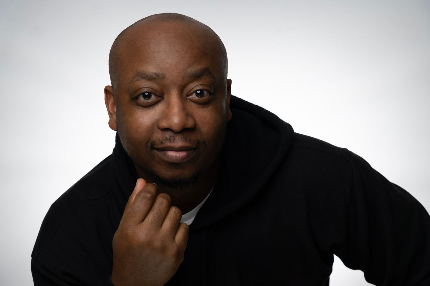Bald Black man in black hoodie poses thoughtfully with hand on chin against gray background.