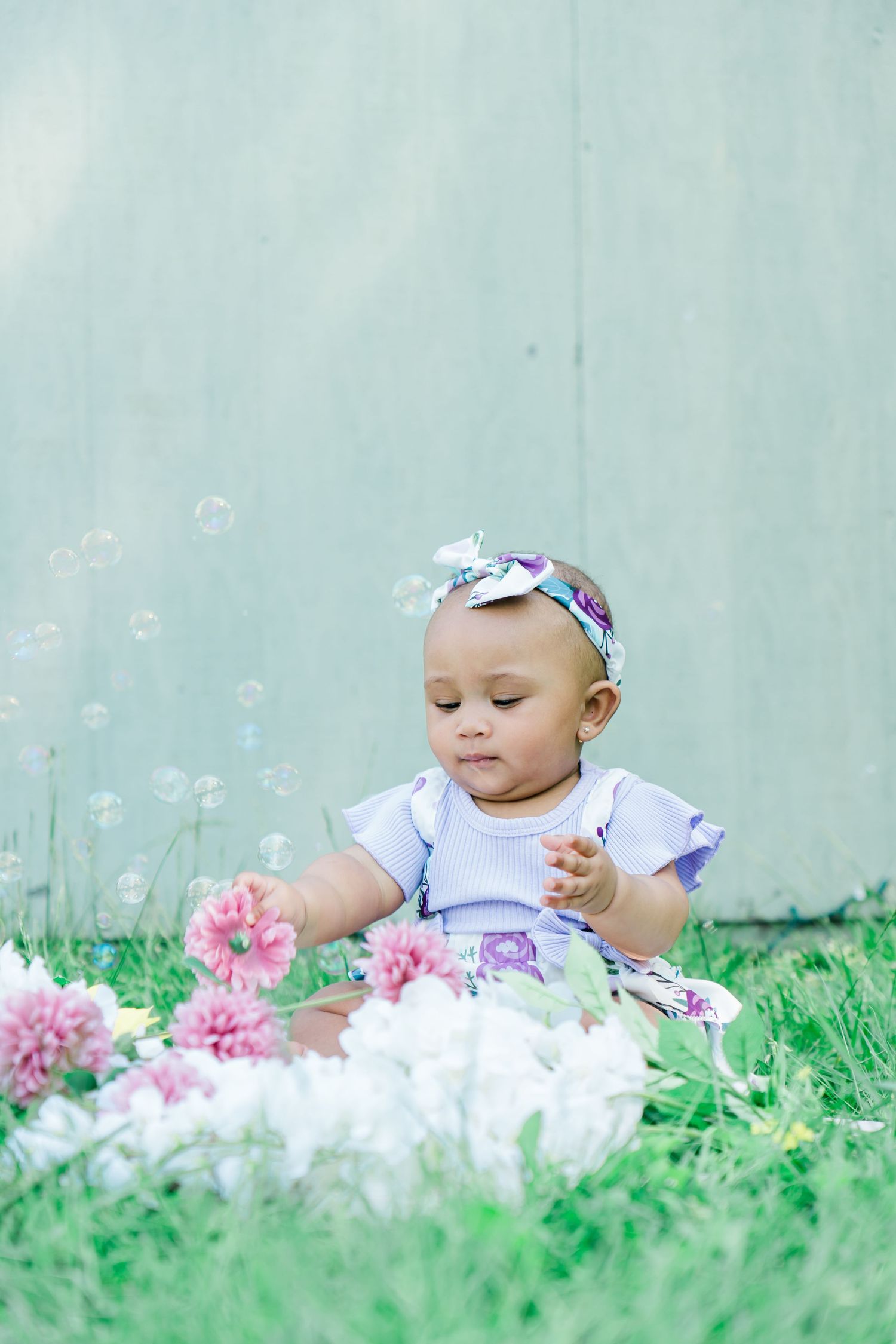 Baby girl in floral dress sitting on grass reaching for pink flowers with bubbles floating nearby.