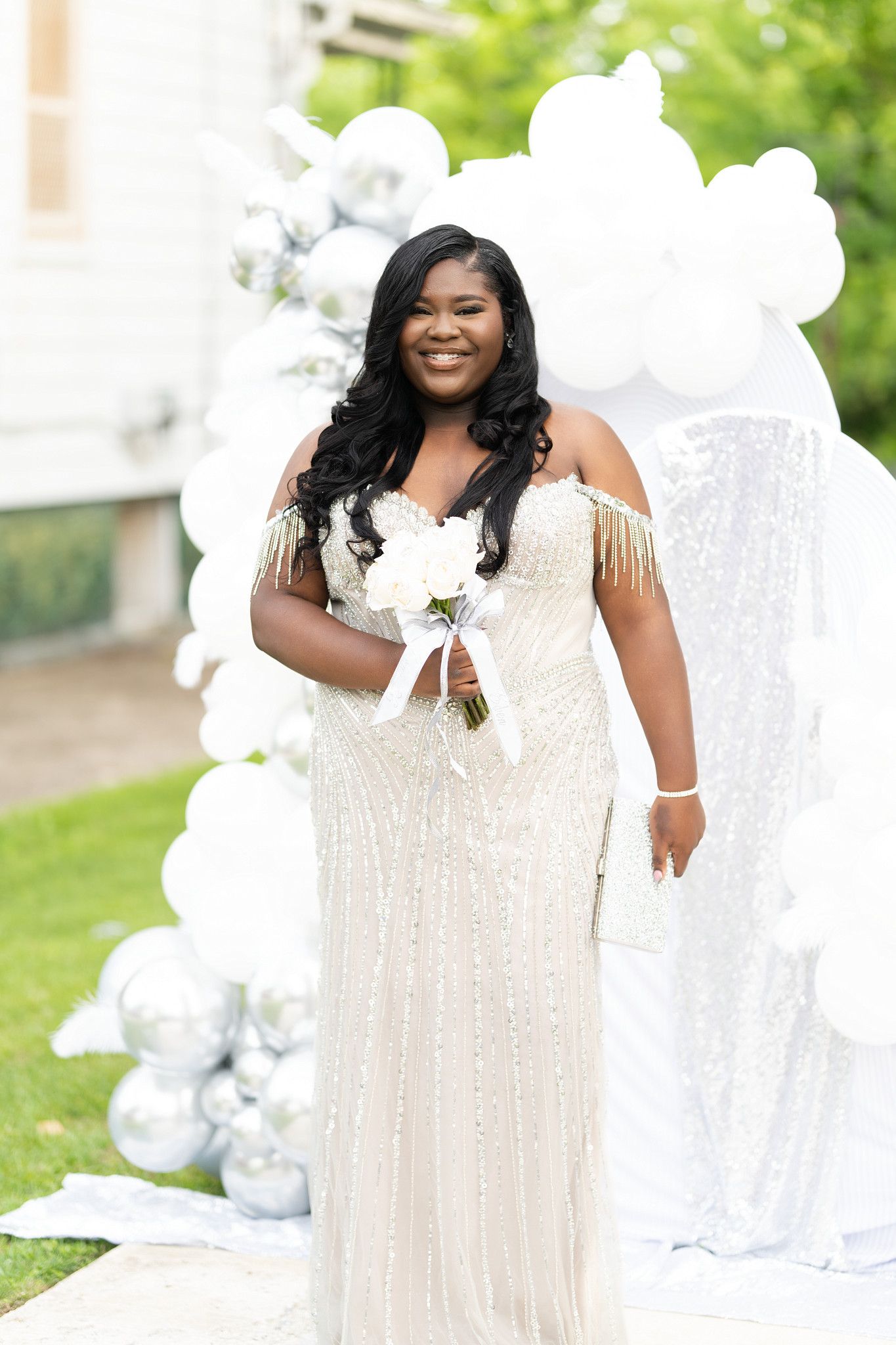 Smiling bride in a beaded off-shoulder white gown holds a bouquet in front of a white balloon arch outdoors.