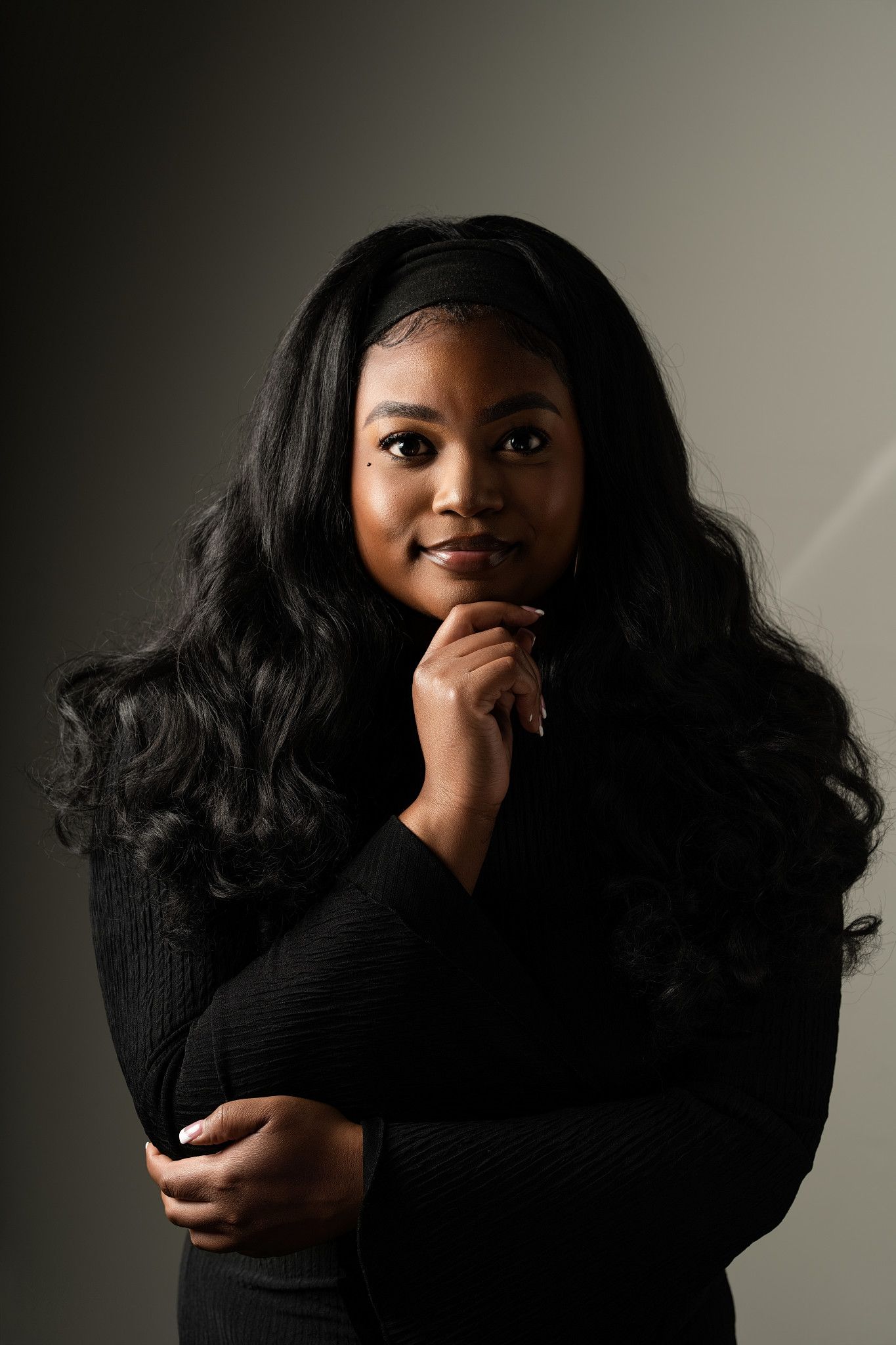 A young Black woman with long wavy hair poses thoughtfully in a black sweater against a dark gray background.