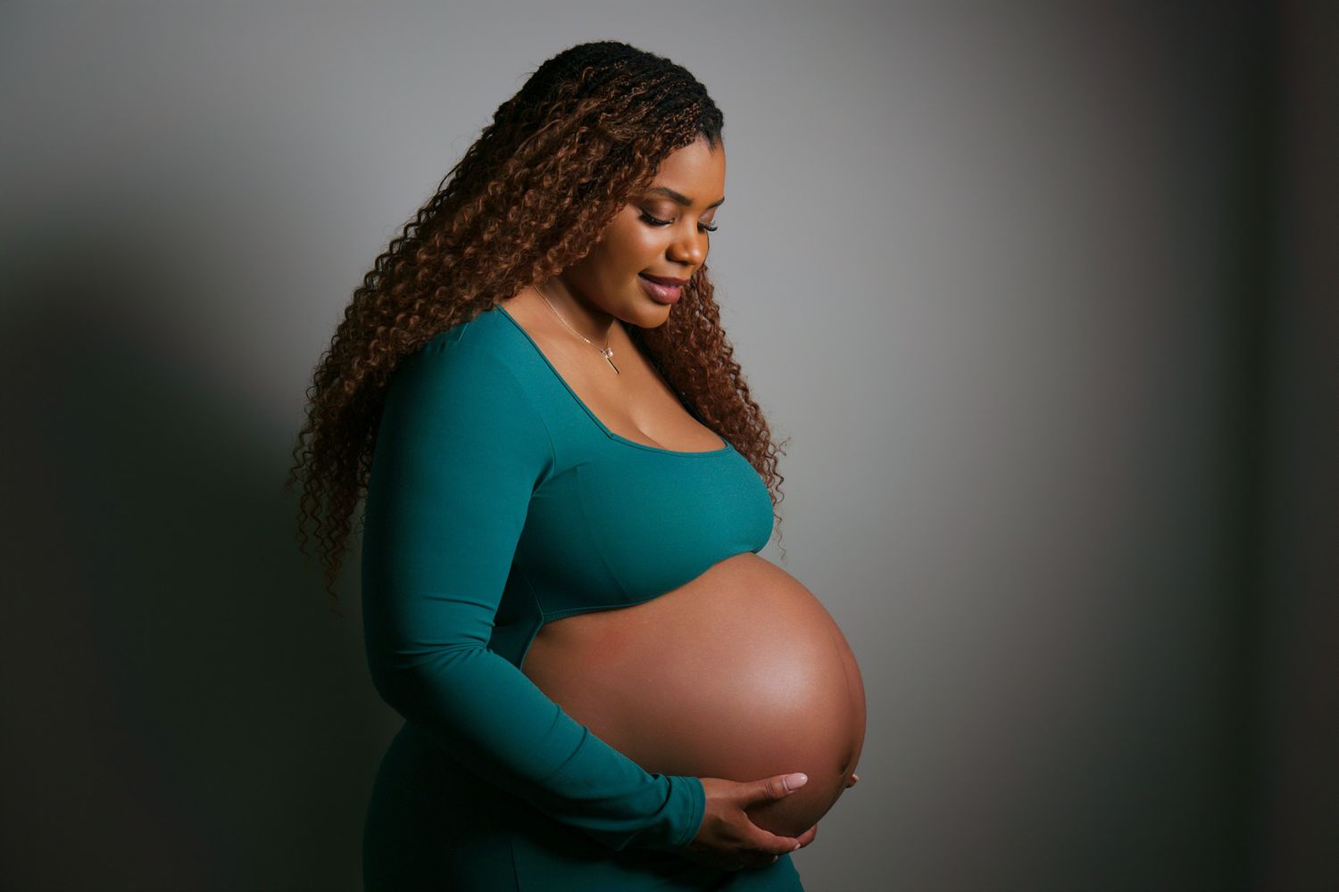 Pregnant Black woman in teal dress cradling baby bump, curly hair, studio portrait with gray background.
