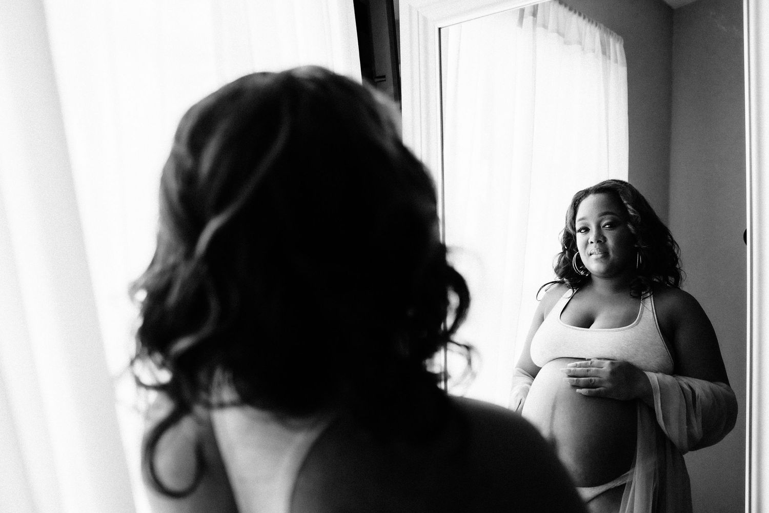 Pregnant woman in lingerie admiring her baby bump in a mirror in black and white photo.
