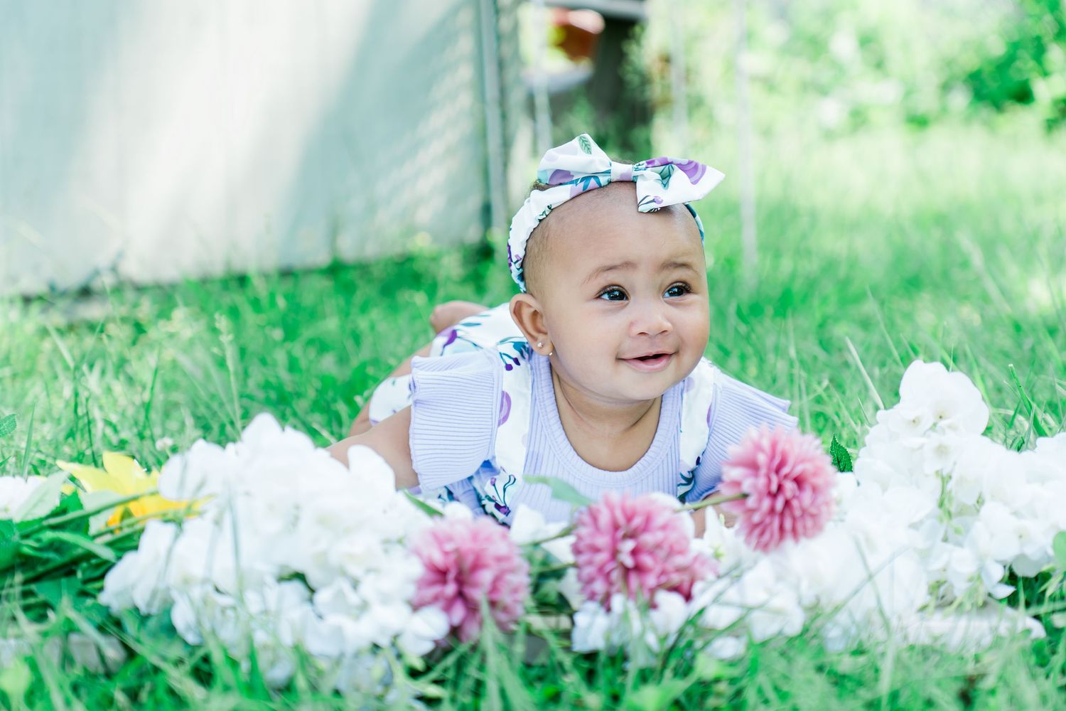 Adorable baby girl with floral headband smiling among white and pink flowers in green grass.
