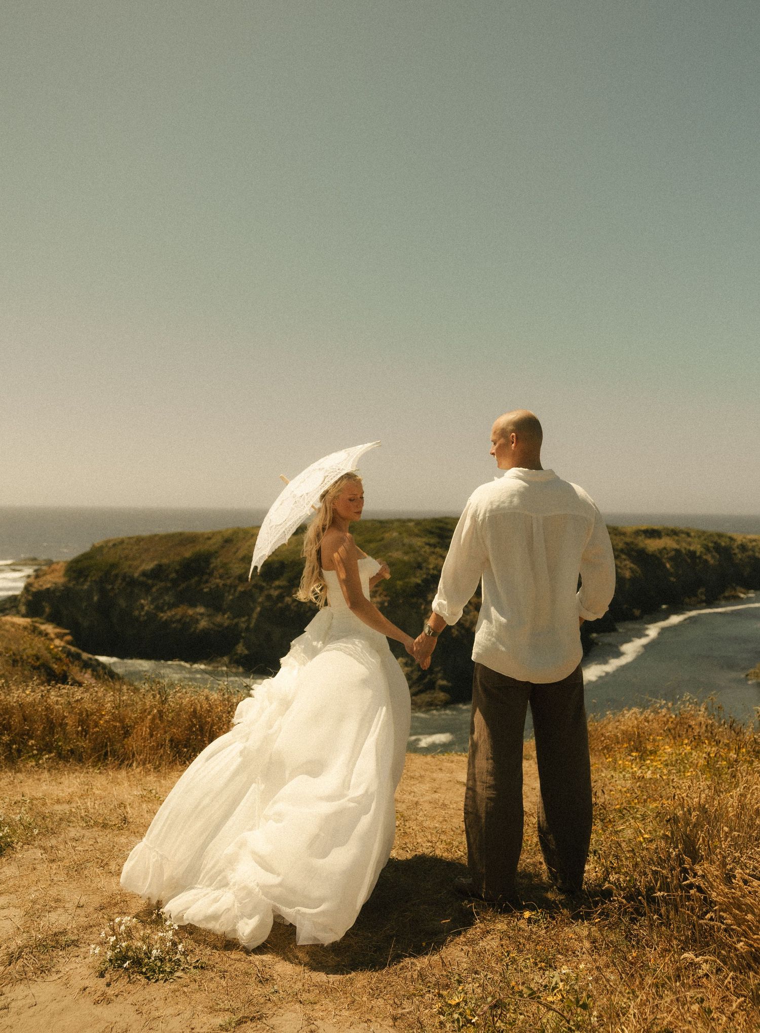 Bride in white gown holding parasol and groom in white shirt hold hands on coastal cliff overlooking ocean.