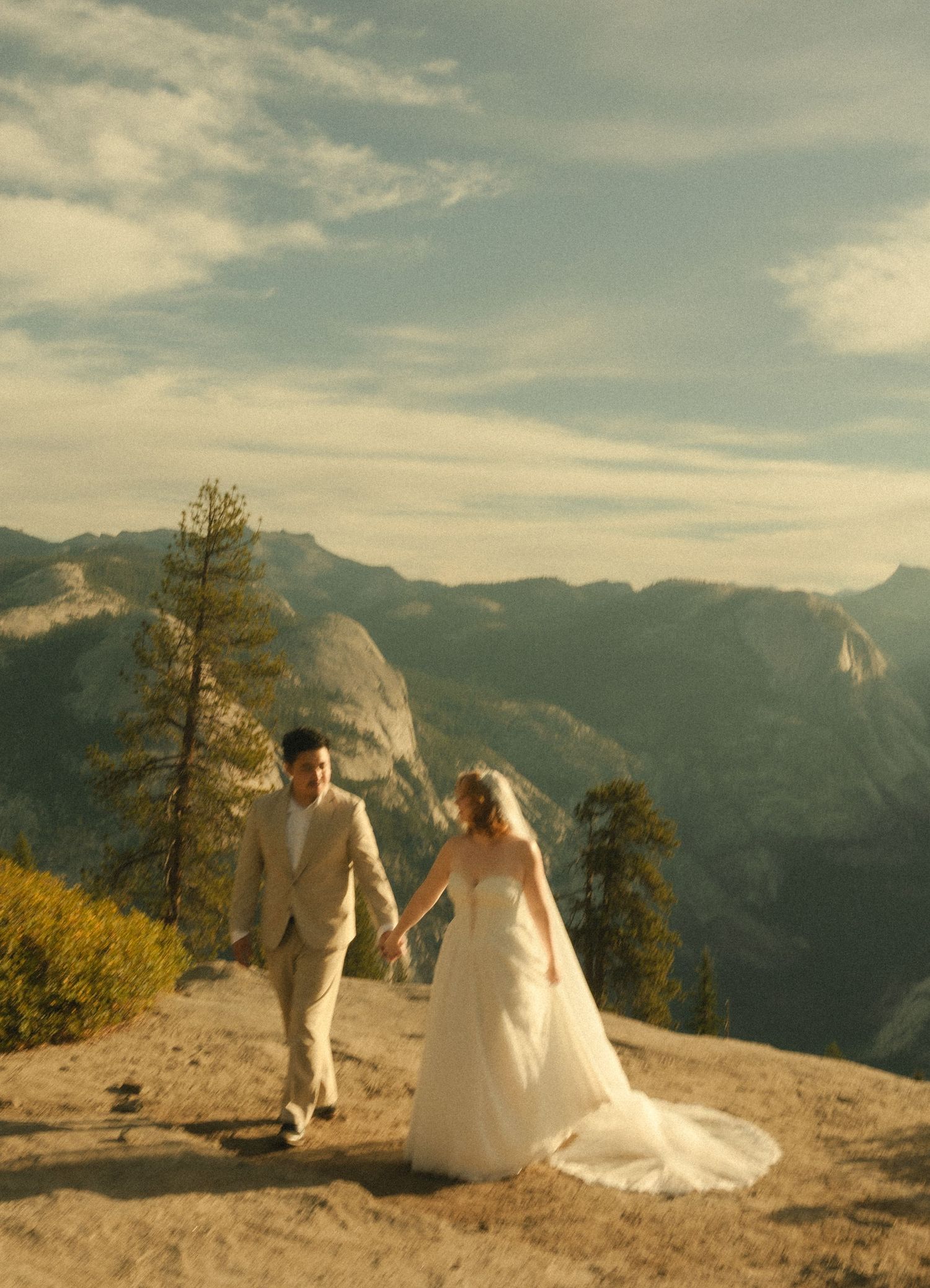 Bride and groom holding hands at Glacier Point, Yosemite, with stunning mountain valley views at golden hour.