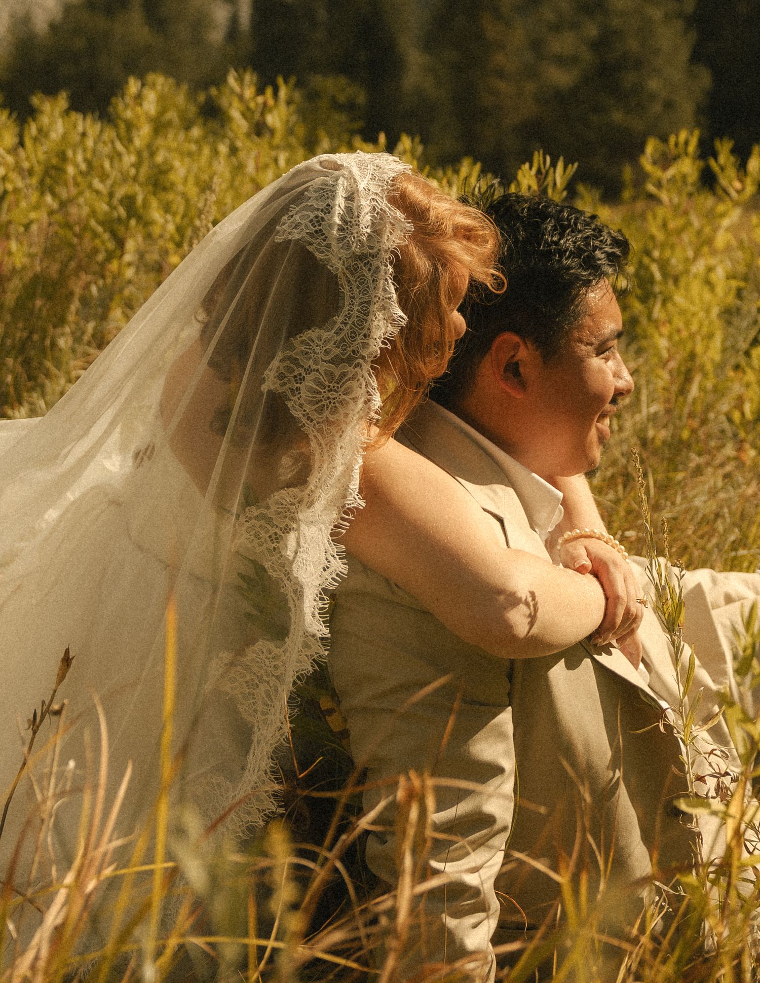 Bride in lace veil embraces groom in tan suit while sitting in golden meadow grass during outdoor wedding.