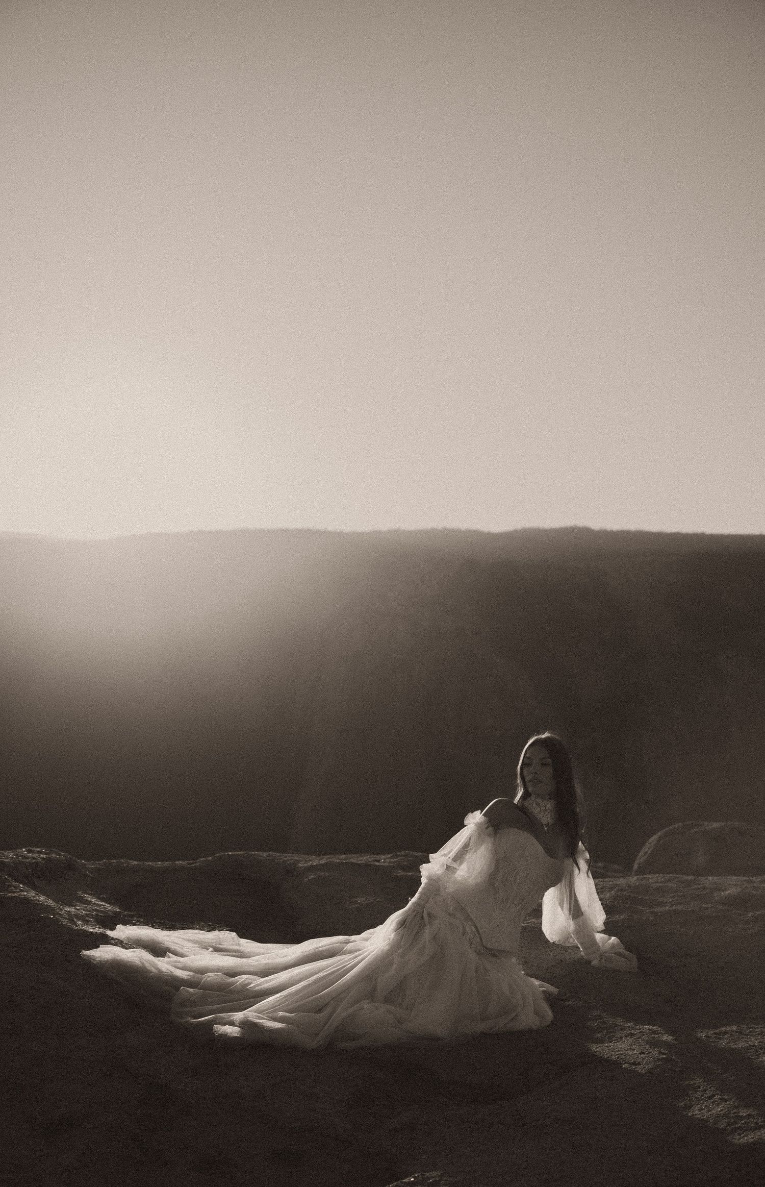 Bride in flowing white gown poses on canyon cliff edge at sunset in dramatic black and white photography.