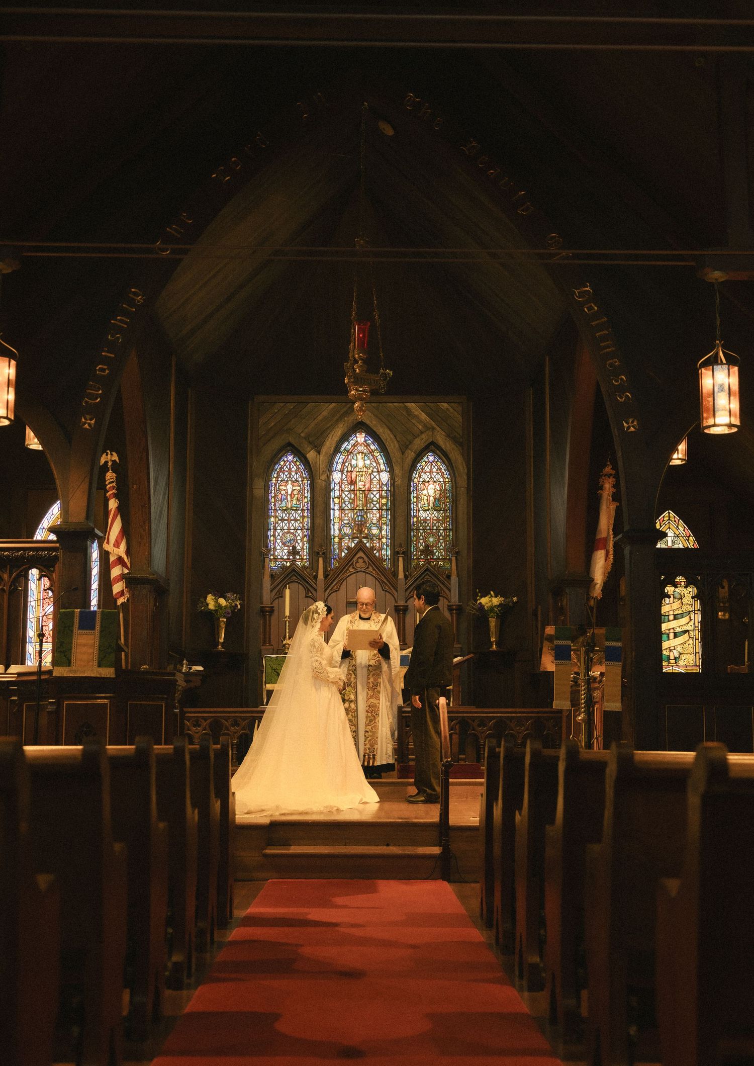 A bride in a white gown stands at the altar of a dark wood church with stained glass windows during a wedding ceremony.