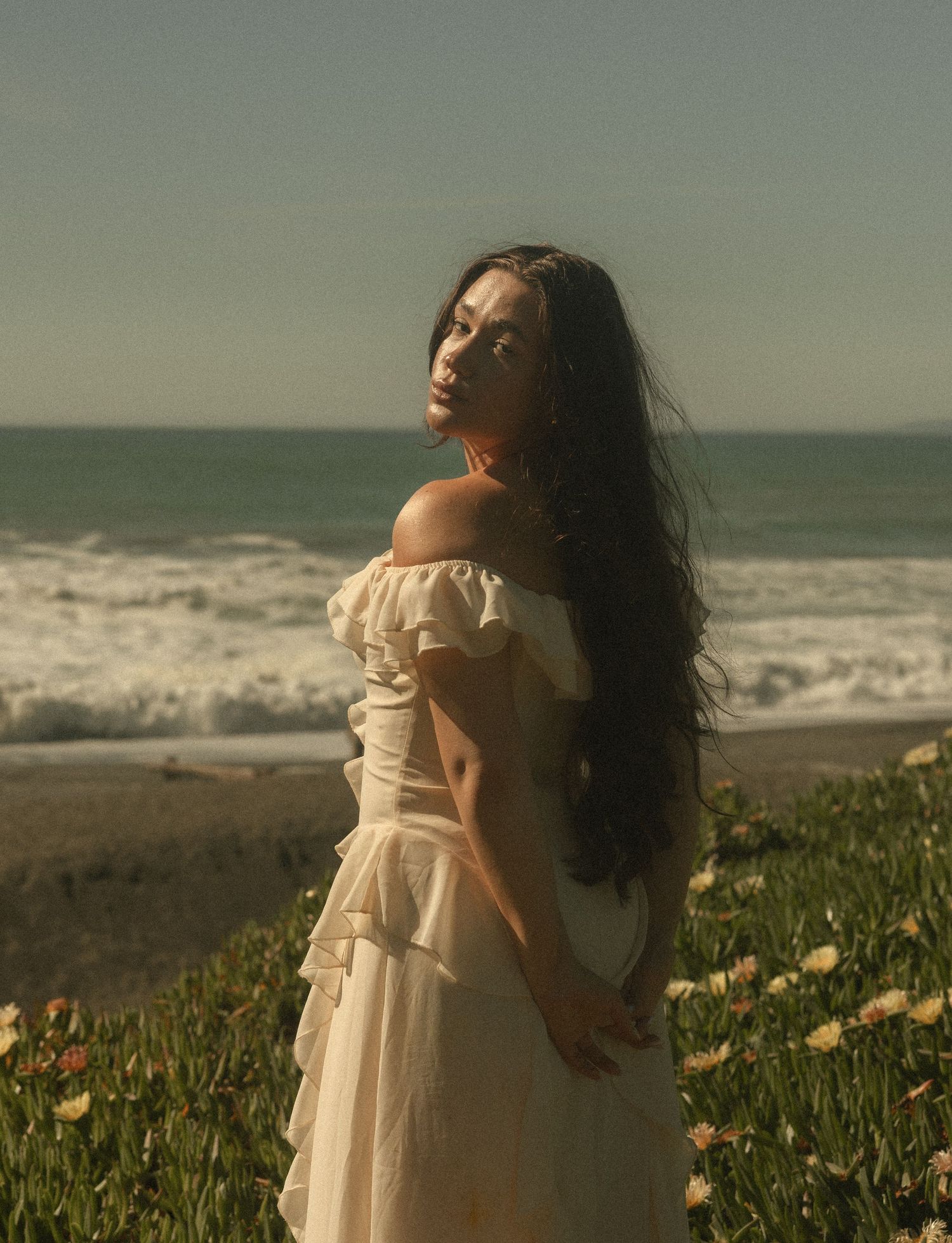 Woman in white off-shoulder dress standing by ocean shore with wildflowers, long dark hair blowing in breeze.