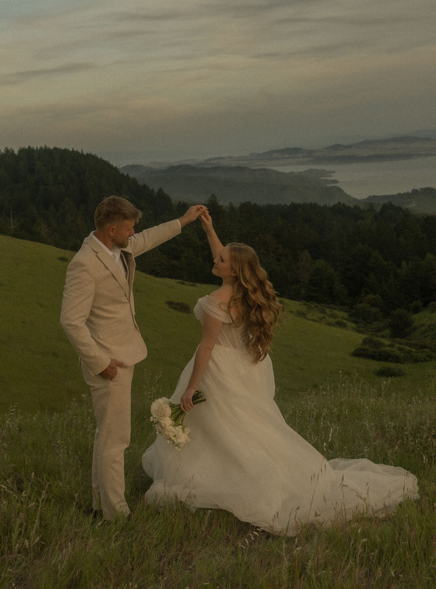 Bride and groom dancing on a scenic hillside meadow at sunset with misty mountains and ocean in the background.