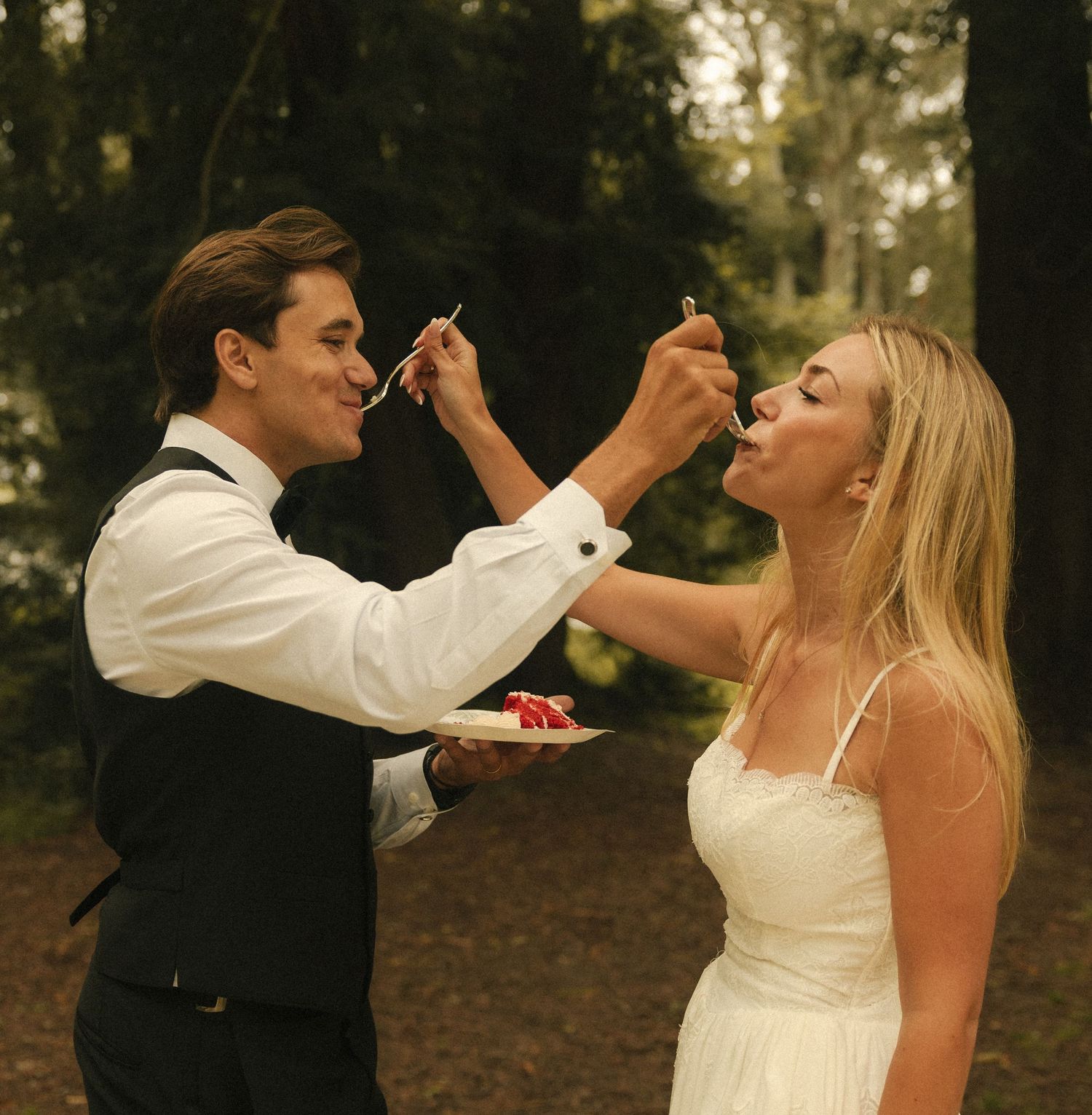 A bride and groom feed each other wedding cake outdoors in a forest setting, both dressed in formal wedding attire.