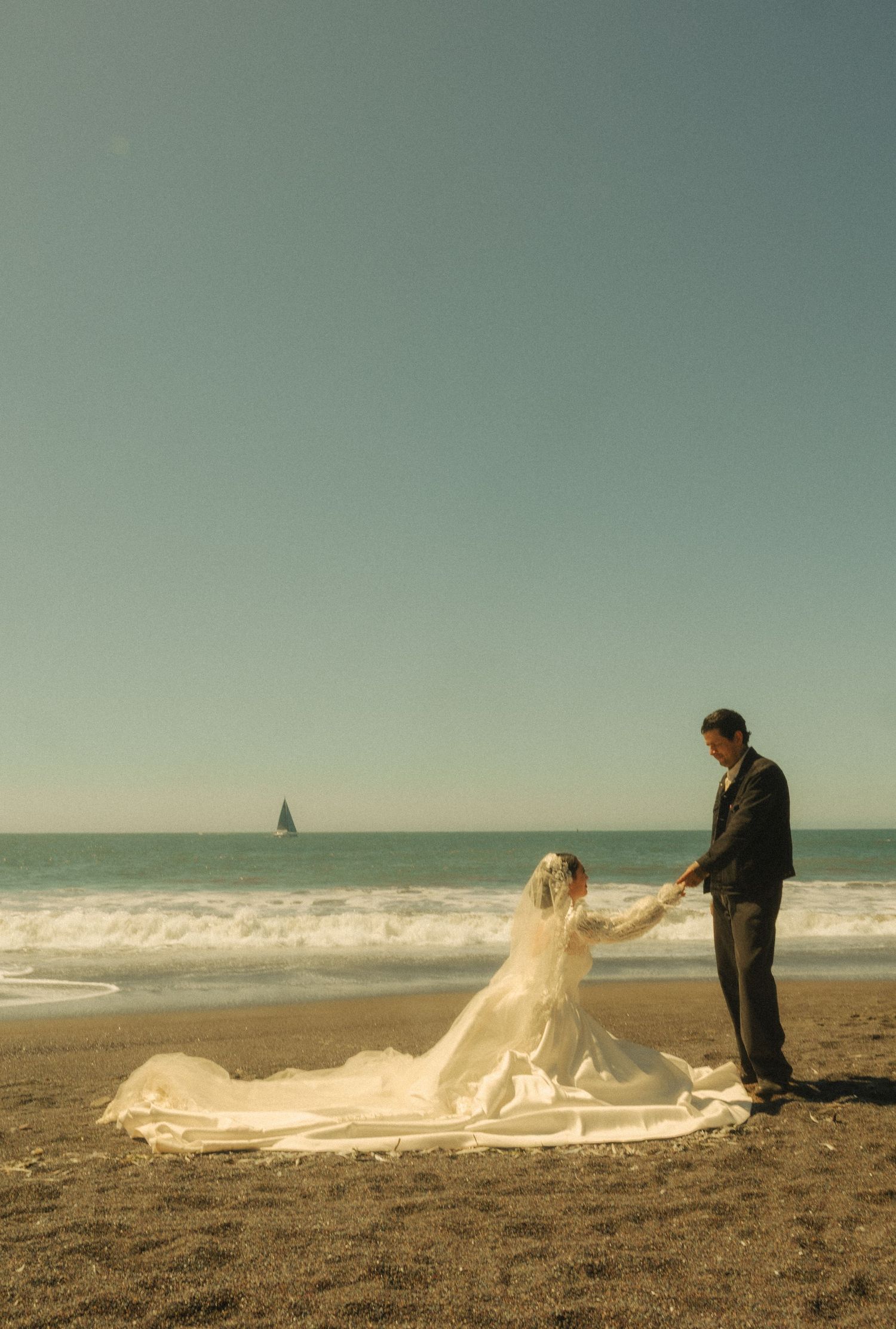 Bride in flowing white gown and veil holds groom's hand on sandy beach with sailboat on horizon.