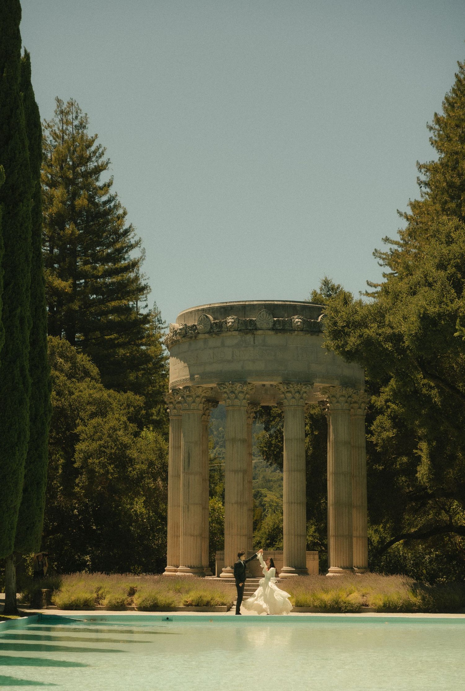 Bride and groom stand before a classical stone rotunda with tall columns, surrounded by lush trees beside a serene pool.
