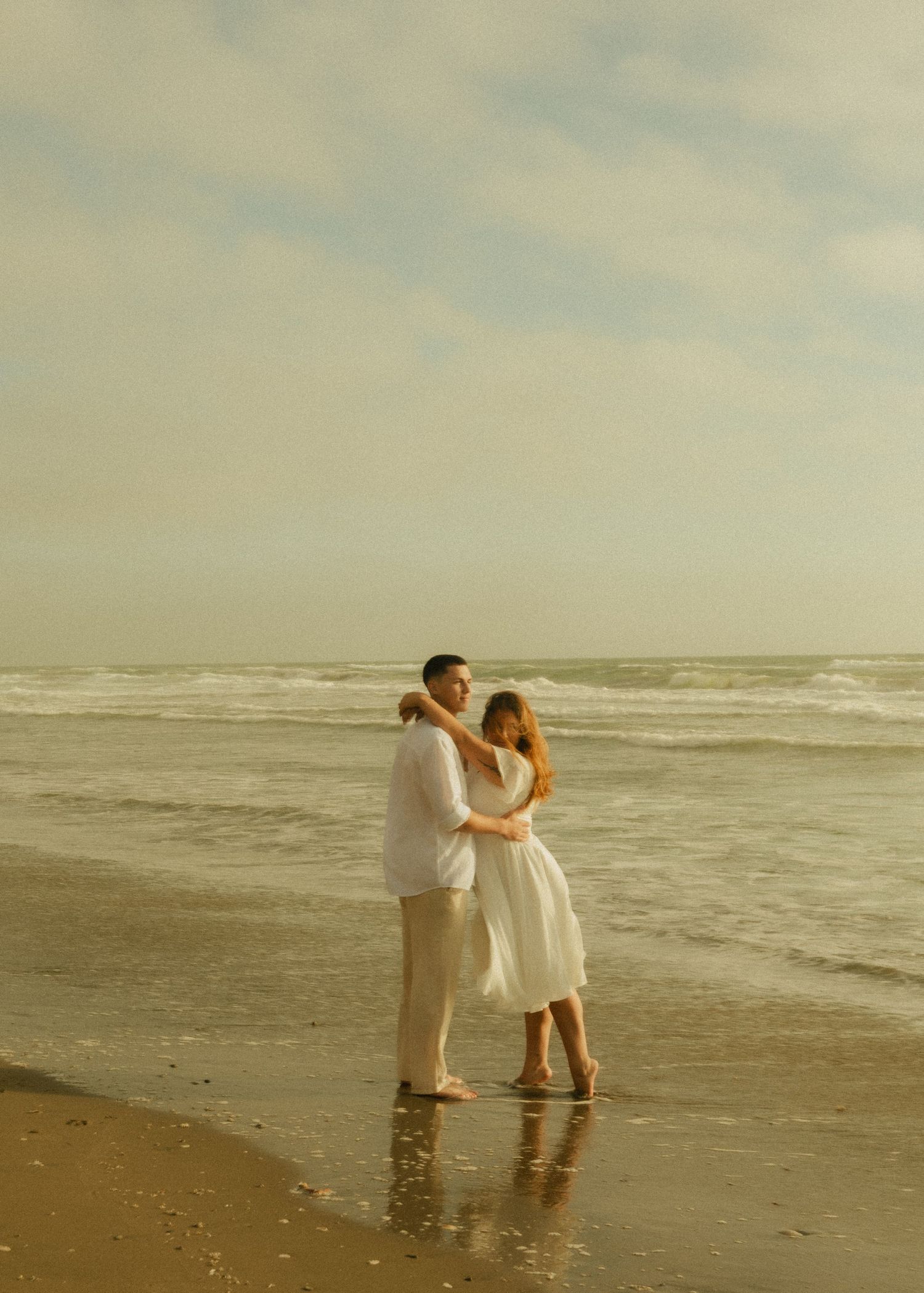 Romantic couple embracing on a sandy beach with ocean waves, dressed in white outfits during golden hour.