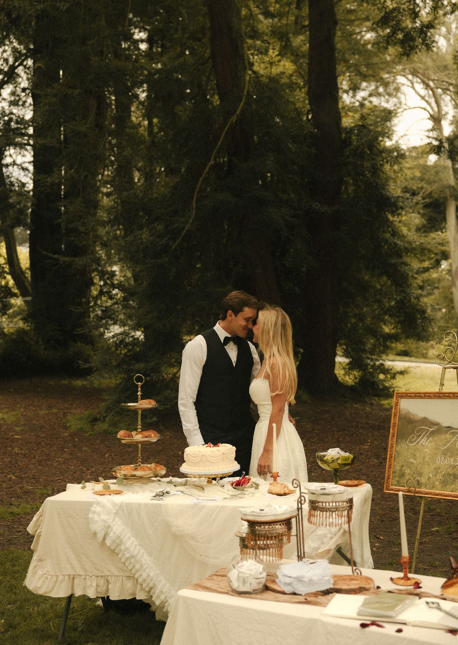Bride and groom sharing a romantic kiss at an outdoor wedding dessert table surrounded by tall trees.