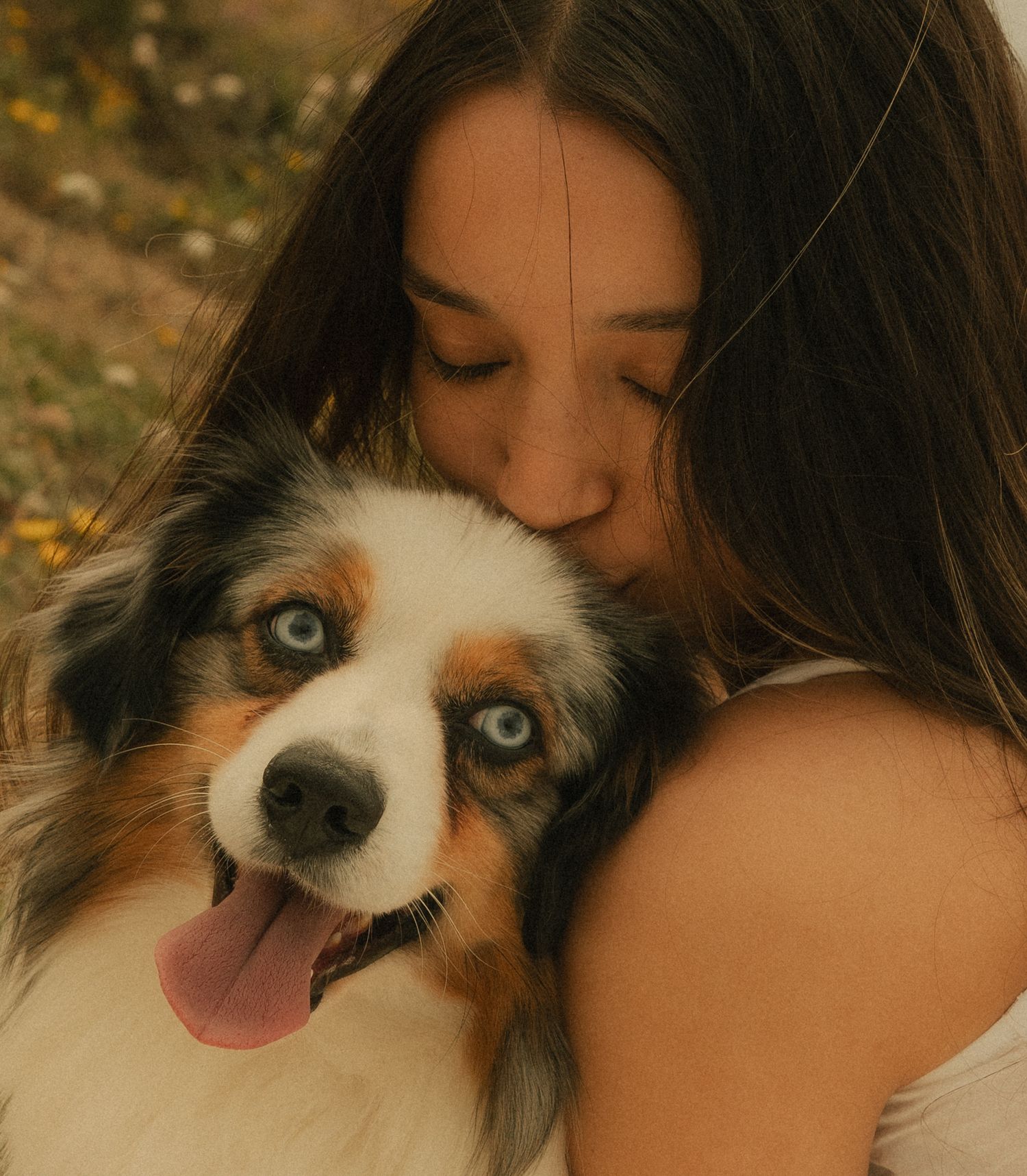 A young woman with long dark hair kisses her blue-eyed Australian Shepherd dog outdoors.