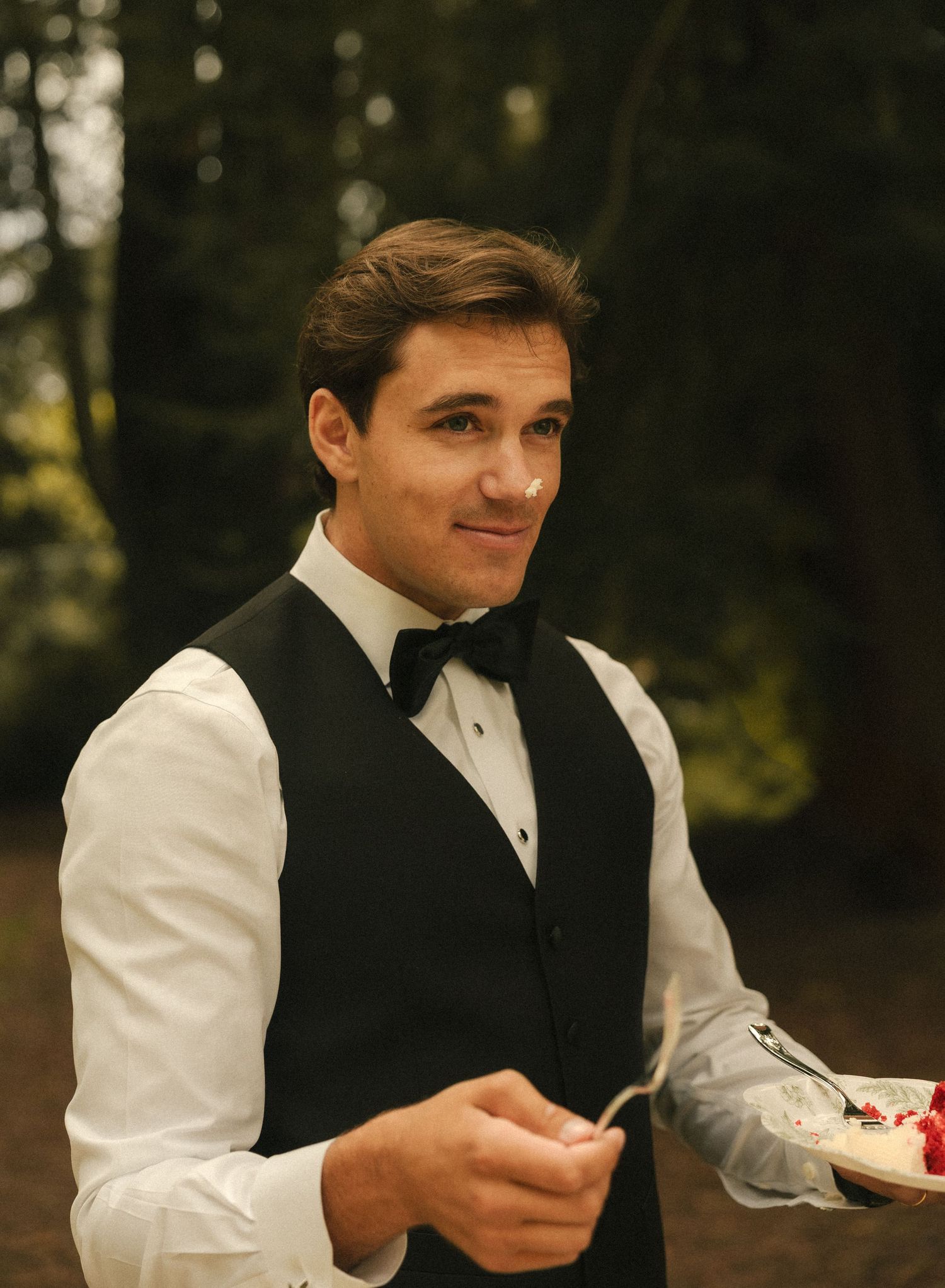 Smiling young man in black vest and bow tie holding a plate of cake outdoors at a formal event.